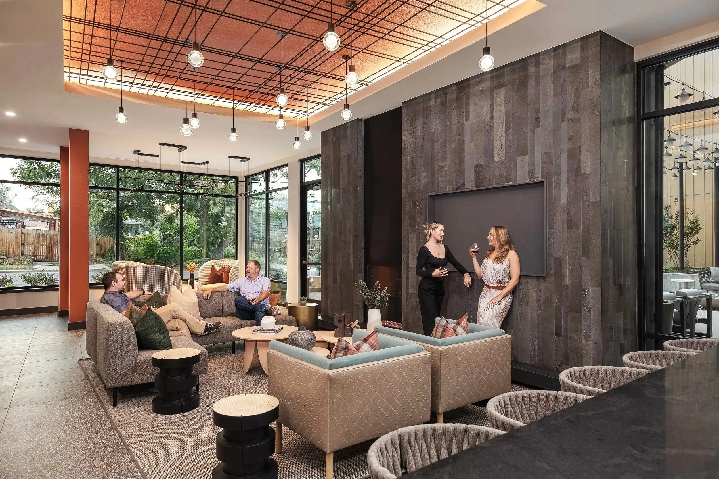 Modern lounge area with large windows, gray sofas, armchairs with patterned cushions, and a dark textured wall with a blank blackboard. Two women are standing and chatting, one holding a glass of wine, while two men are lounging on the sofas.