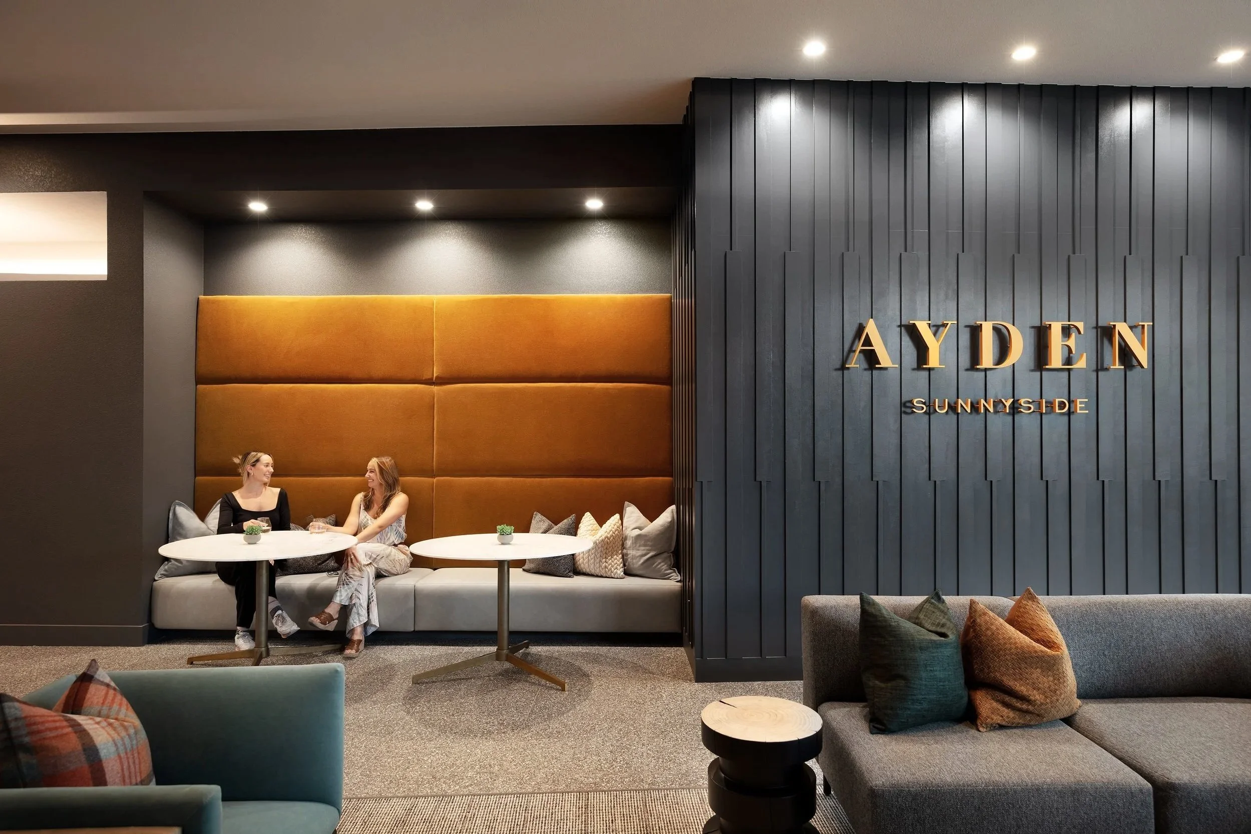 Two women sitting and talking at a round table in a modern hotel lounge with a dark gray and blue color scheme, gold signage reading 'AYDEN SUNNY SIDE,' and various decorative pillows and furniture.
