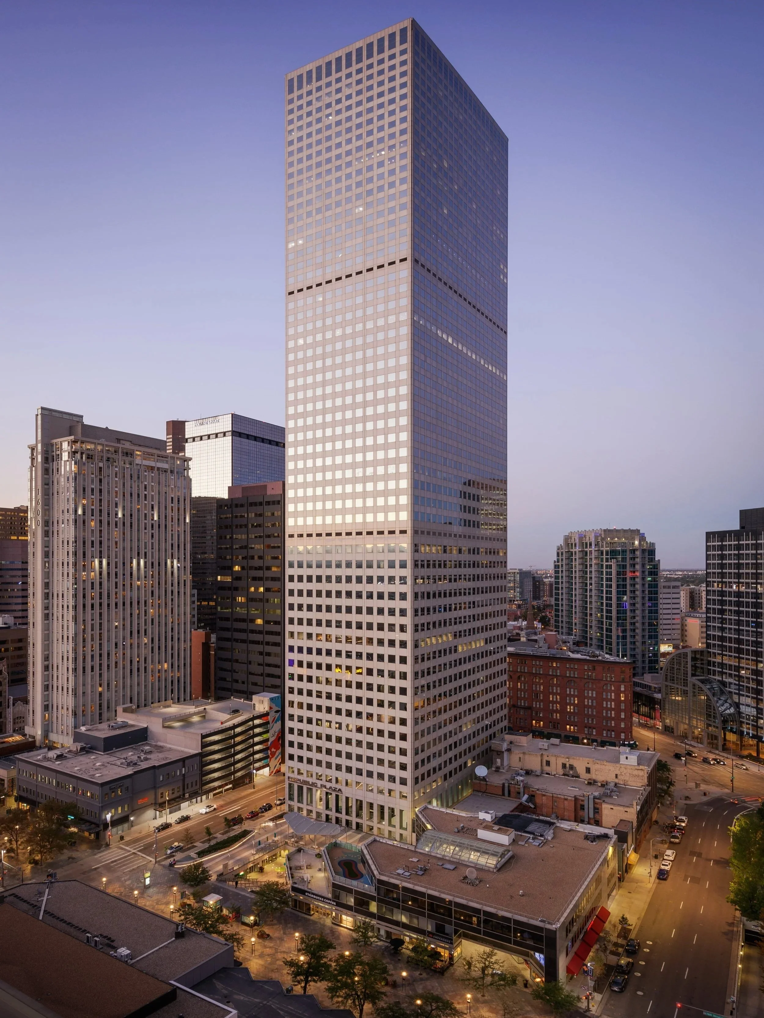 Tall modern office building in downtown cityscape during twilight, surrounded by other high-rise buildings and street level activity.