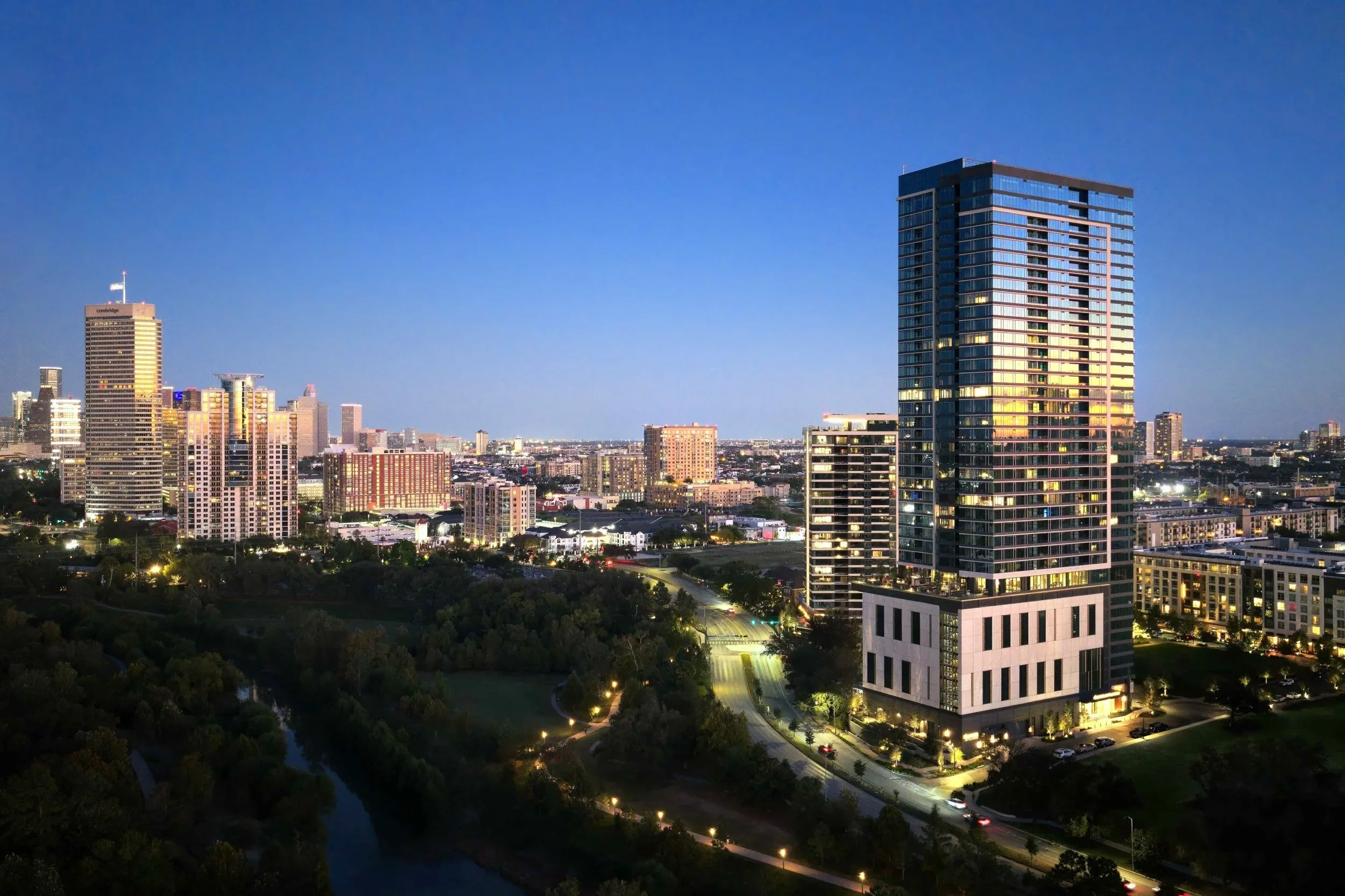 A city skyline at dusk with illuminated high-rise buildings, a river in the foreground, and a clear darkening sky.