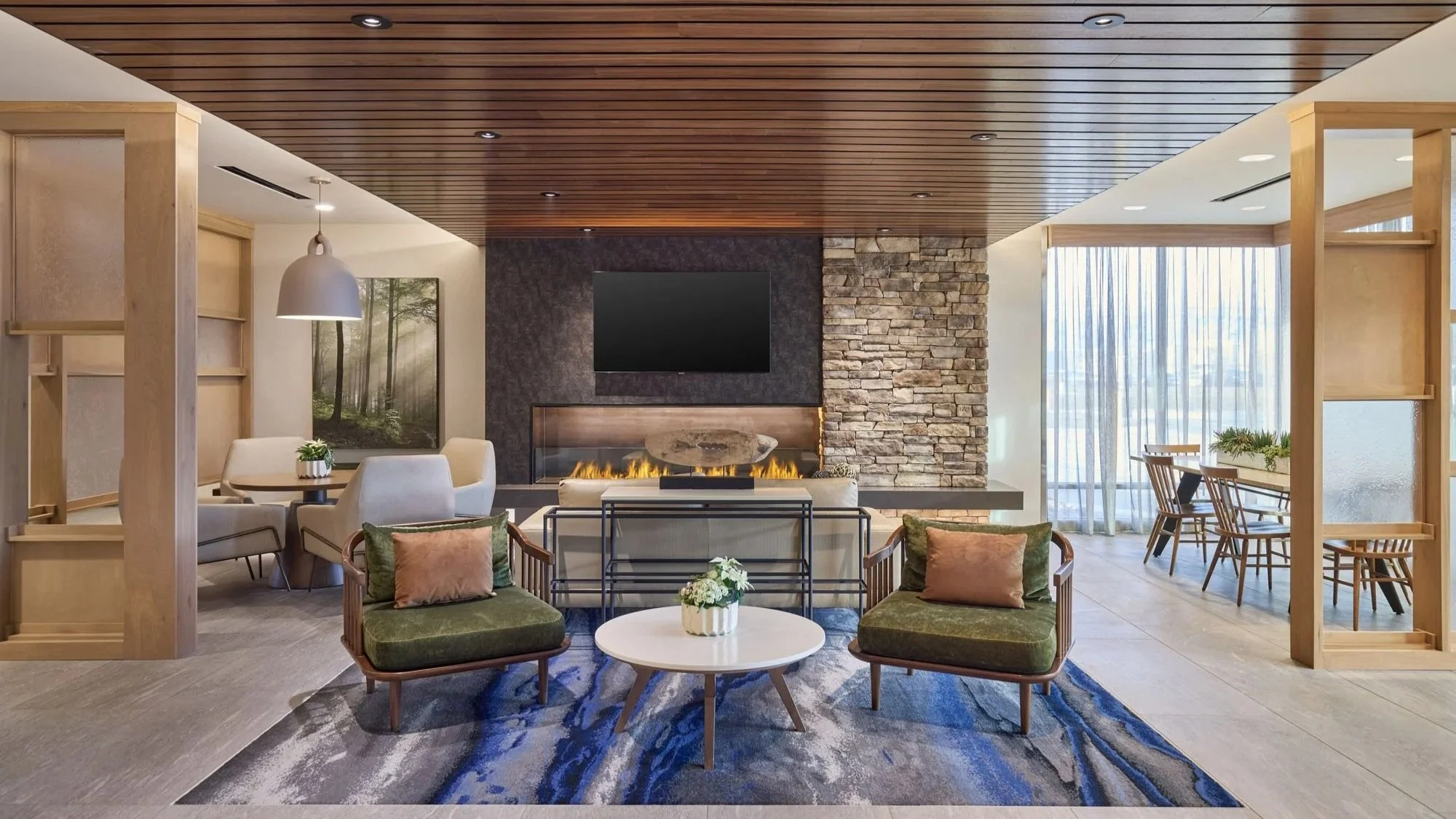 Modern living room with a fireplace, wall-mounted TV, wooden ceiling, green armchairs, white round coffee table with flowers, shared dining area, and large windows with sheer curtains.