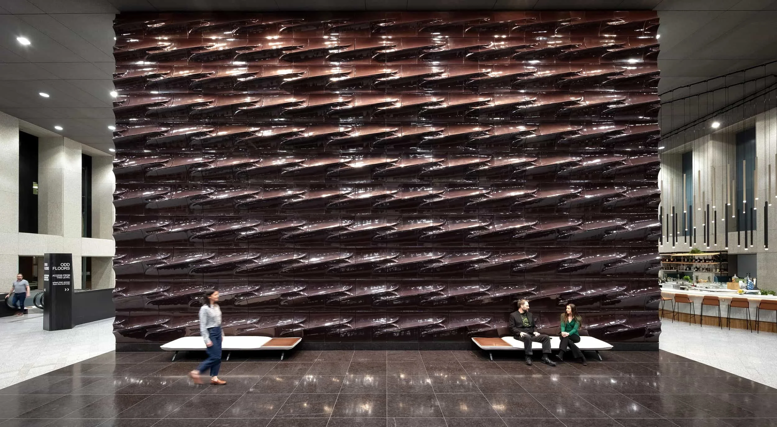 Interior of a modern building with a large abstract dark colored wall art, two people sitting on benches near the wall, and another person walking past.