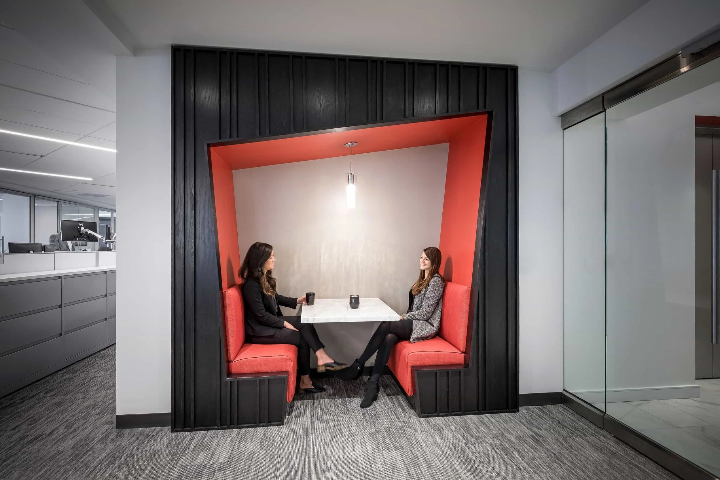 Two women sitting in a modern, enclosed booth with red cushioned seats and a white table, having a conversation in an office setting.