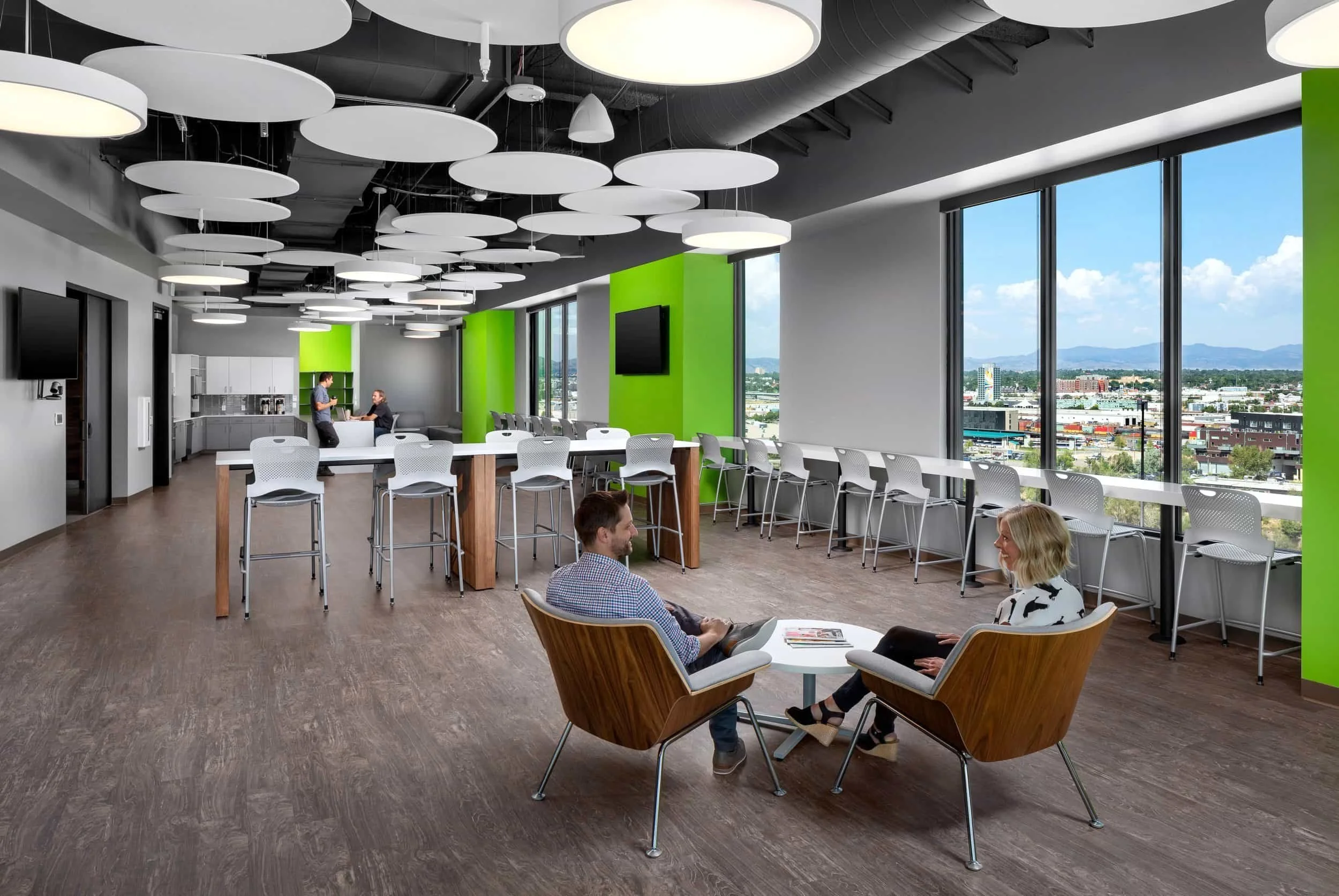 Modern office lounge with large windows, white chairs, and two people talking in chairs around a small table.