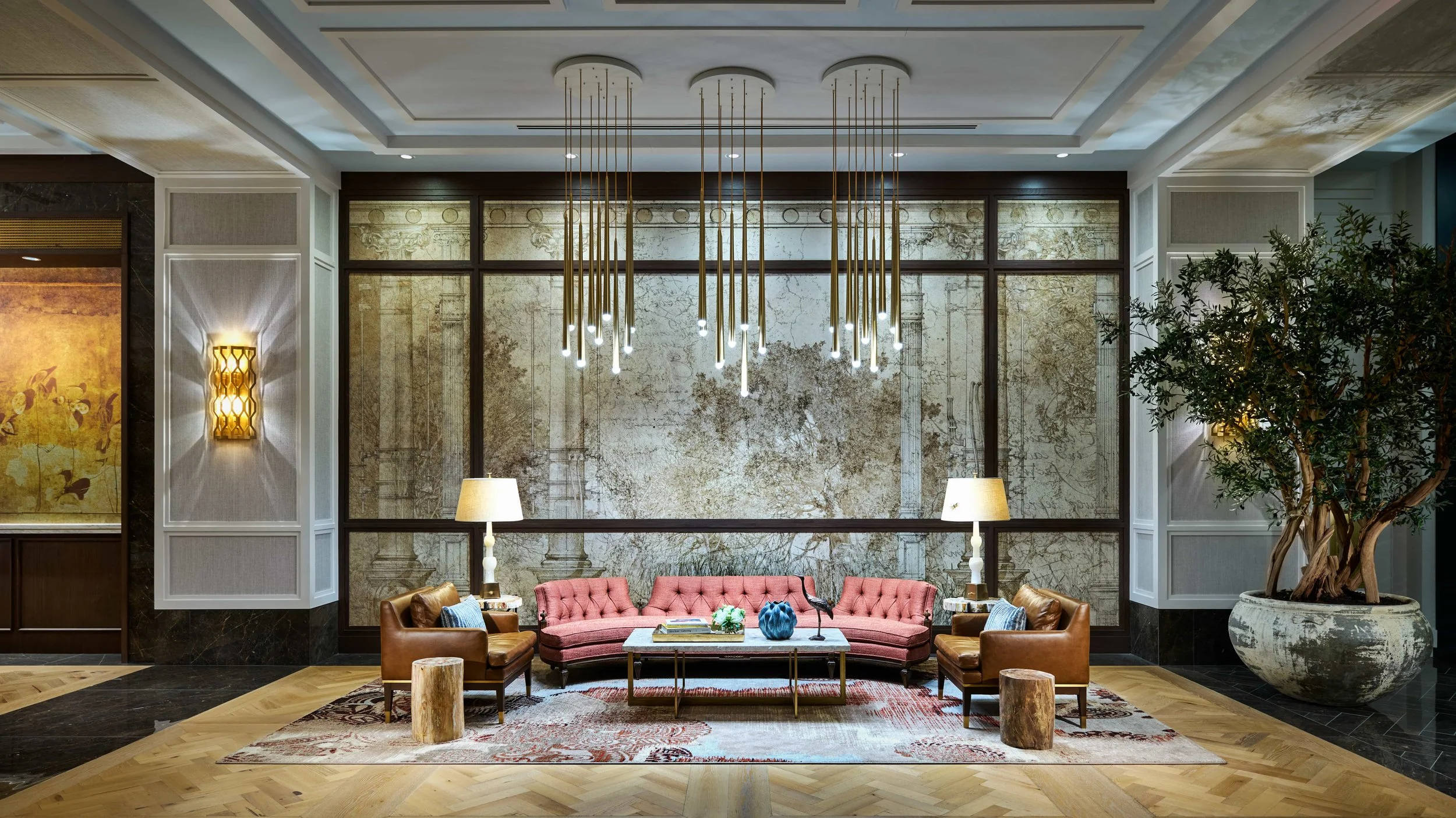 A modern hotel lobby with a pink velvet sofa, two armchairs, two lamps, a coffee table with decorative items, and a large potted plant. Overhead are gold light fixtures hanging from the ceiling. The backdrop features a textured wall with metallic acc