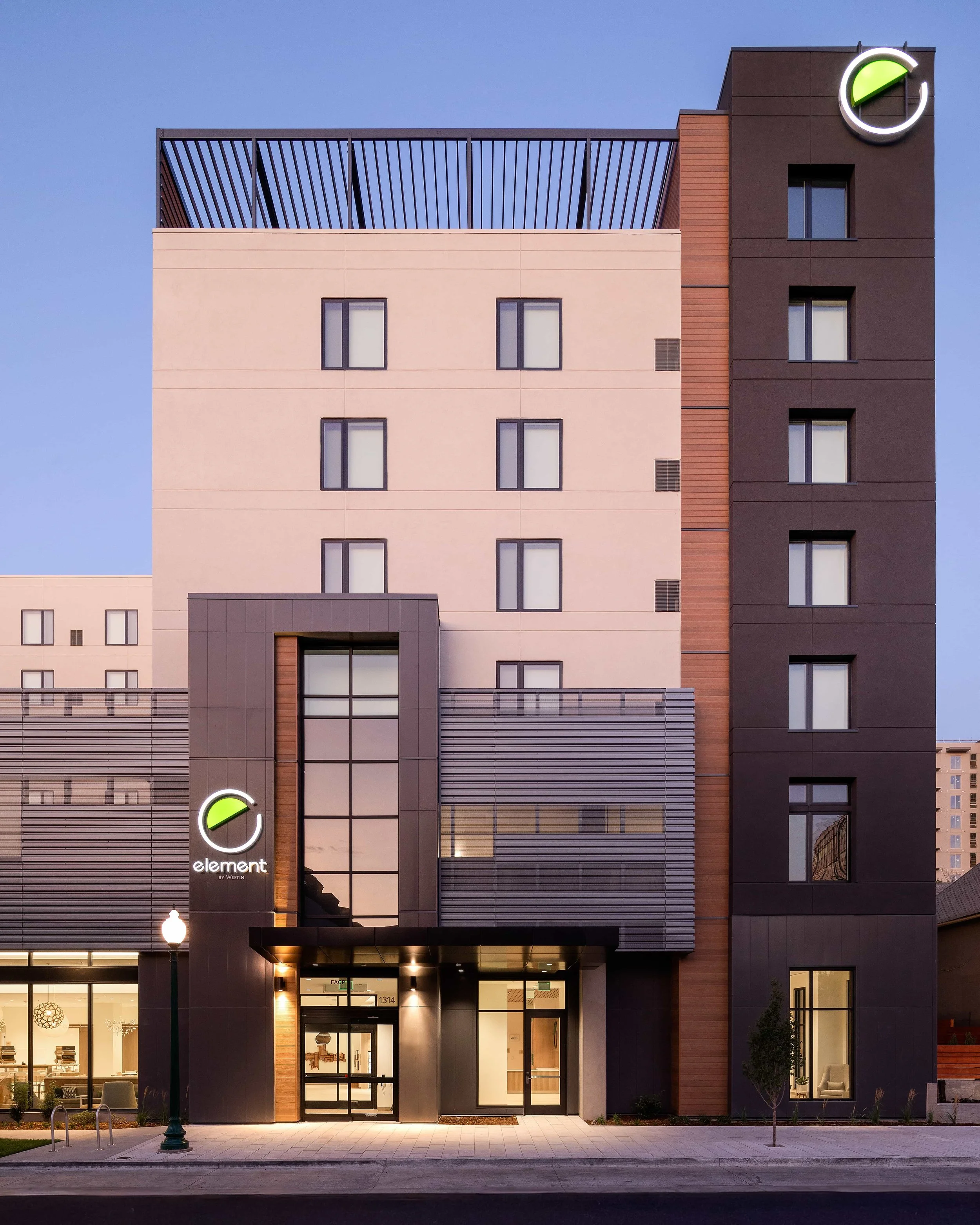 Modern multi-story building with signage that says 'element by Westin', illuminated at dusk with lit entrance, large windows, and a sidewalk with a streetlamp and small tree in front.