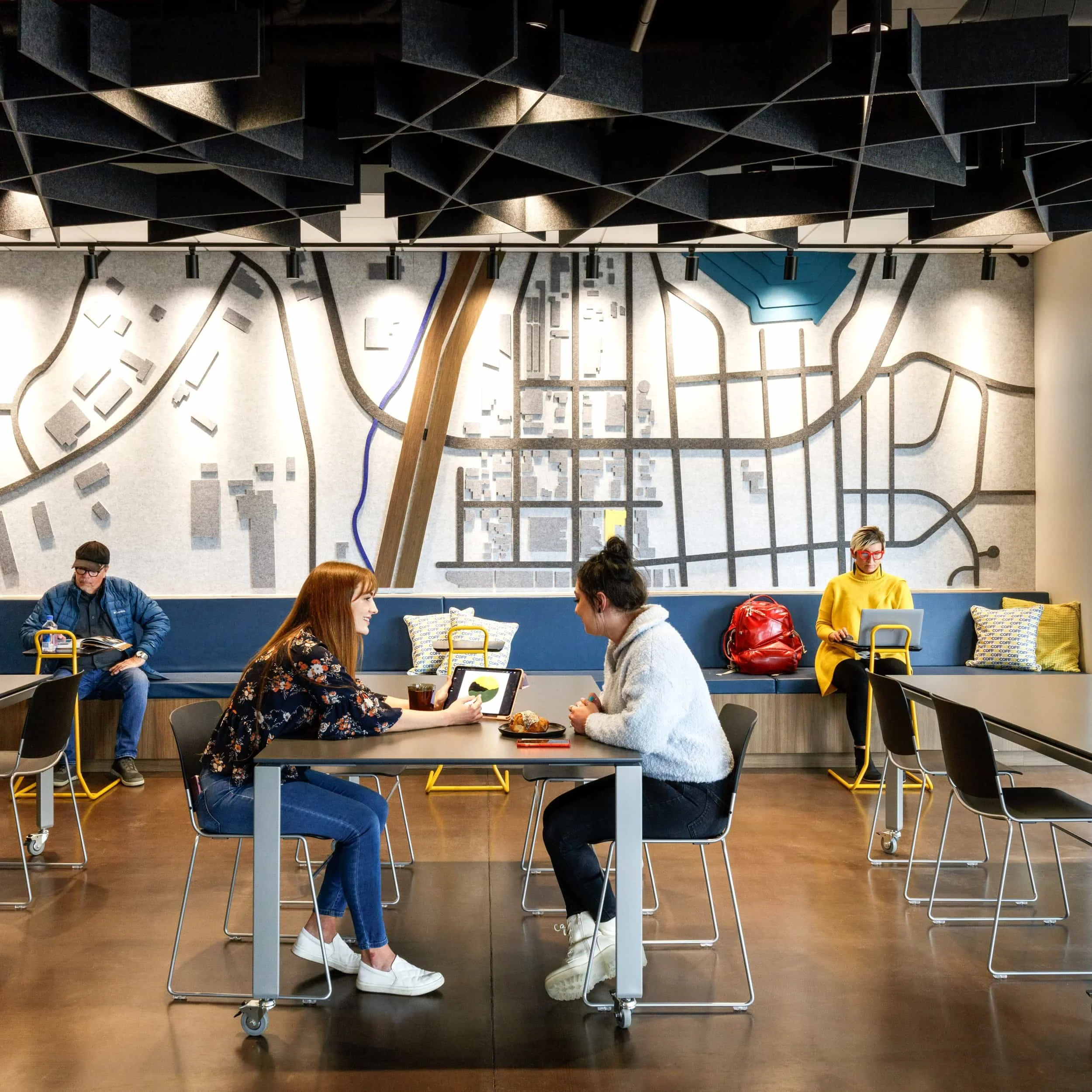 Two women sitting at a table in a modern cafe, engaged in conversation, with one showing something on her tablet. In the background, a man reads a book on a bench to the left, and a woman in a yellow sweater, with red glasses and a red backpack, sits