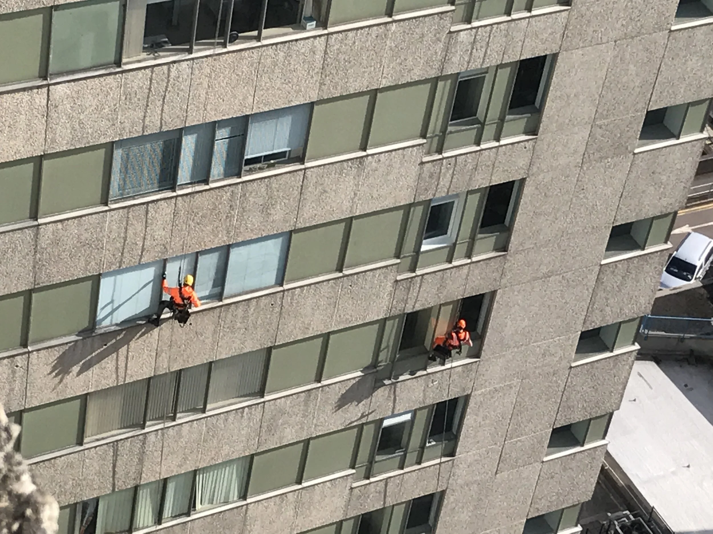 Two window washers in orange uniforms and helmets cleaning the windows of a high-rise building.