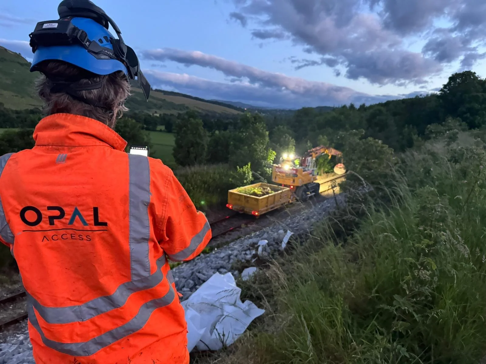 A person wearing an orange safety jacket and a blue helmet, standing beside a railway track at dusk, observing a locomotive train that has derailed and is resting among trees and bushes in a green, hilly landscape.