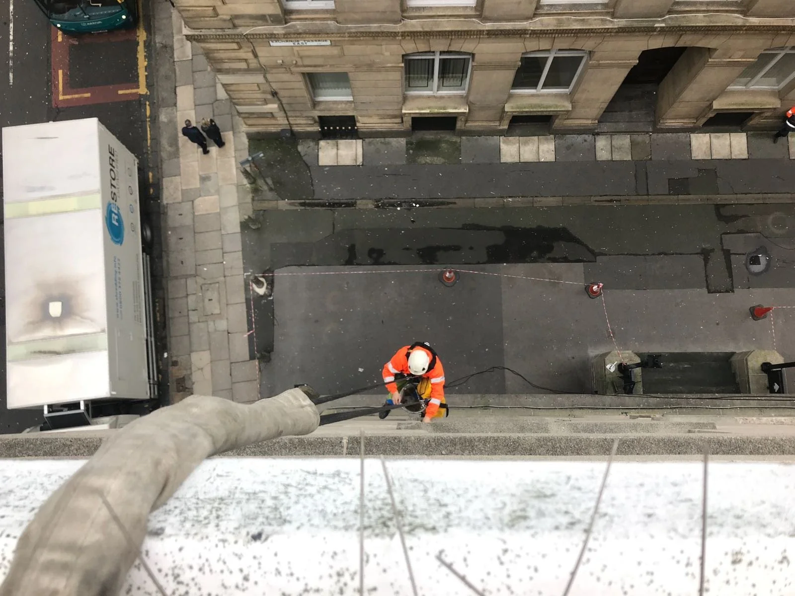 Worker in safety gear cleaning the exterior of a high-rise building with a power washer, viewed from above.