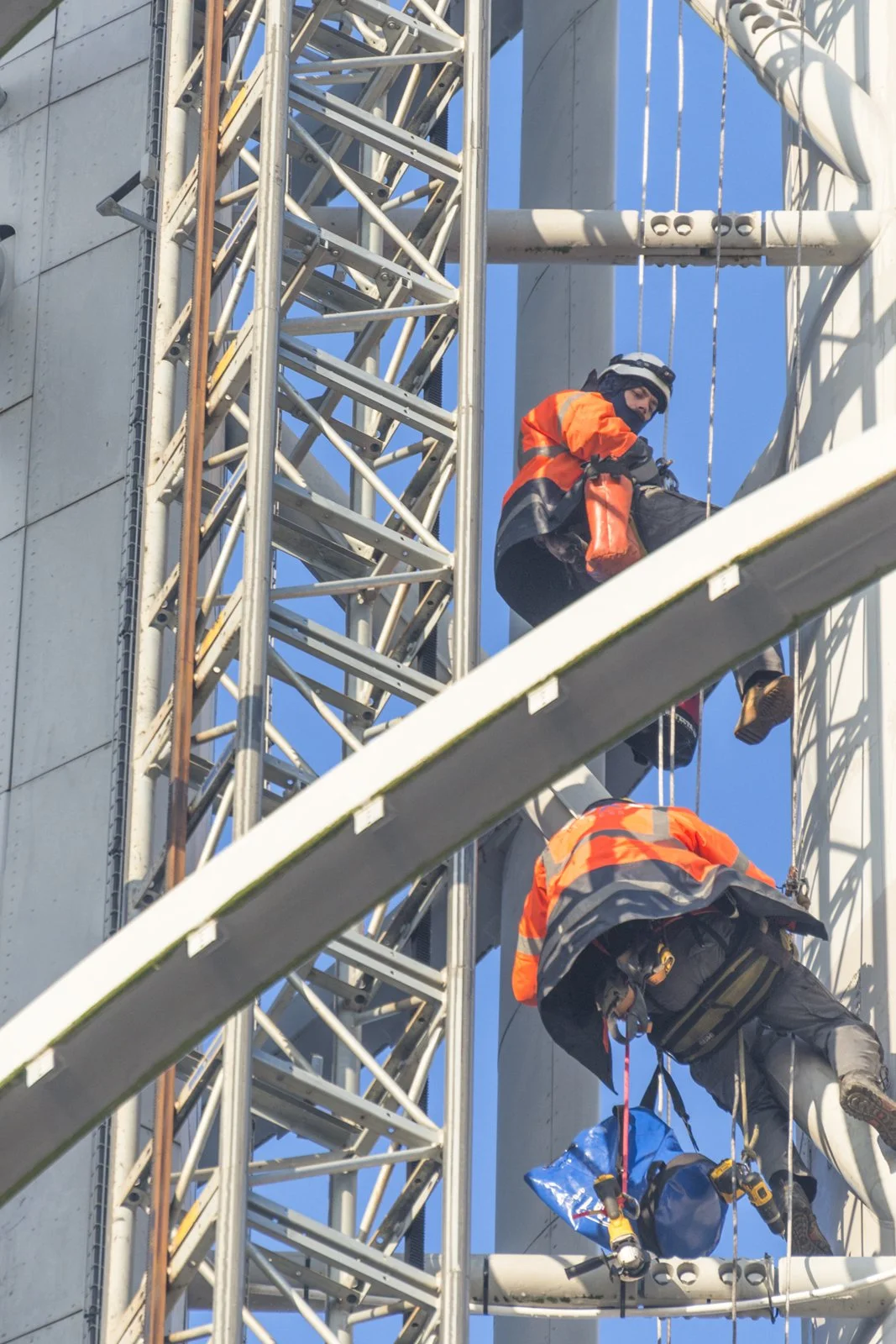 Two construction workers wearing orange safety vests and helmets working on a tall metal building or structure, with one worker climbing and the other sitting on a platform.