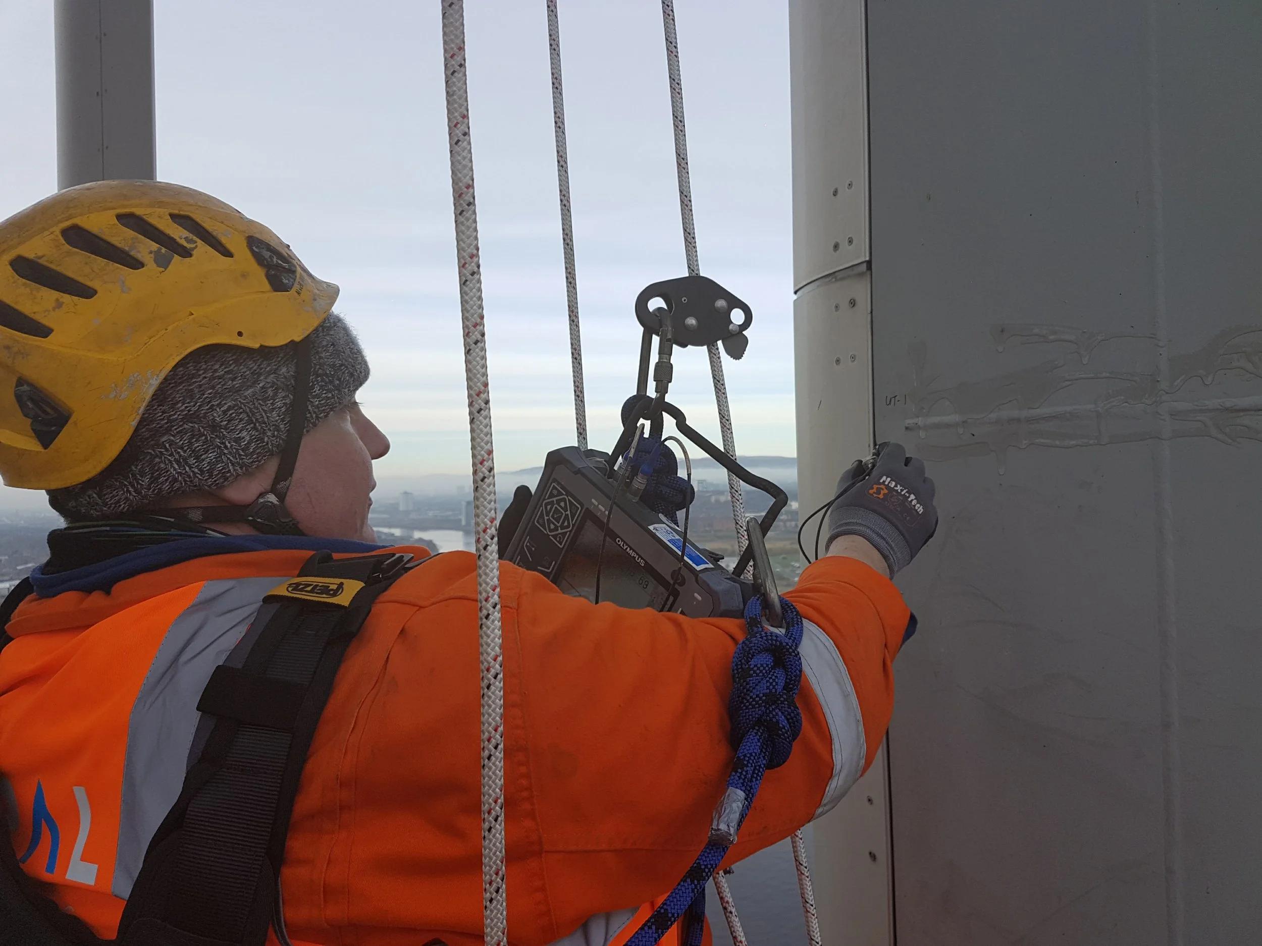 A worker wearing a yellow safety helmet and orange jacket inspecting or repairing a structure high above the ground, with a cityscape and body of water visible in the background.