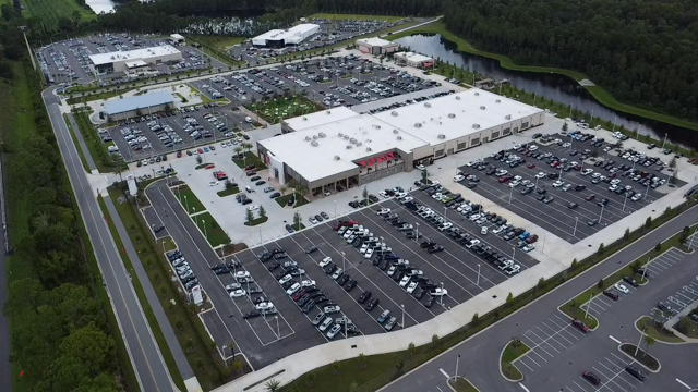 Aerial view of a large shopping center with parking lots and surrounded by greenery, with a body of water nearby.