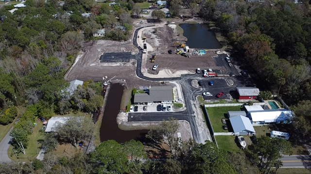 Aerial view of a construction site next to a small pond and residential houses, with some vehicles and construction equipment.