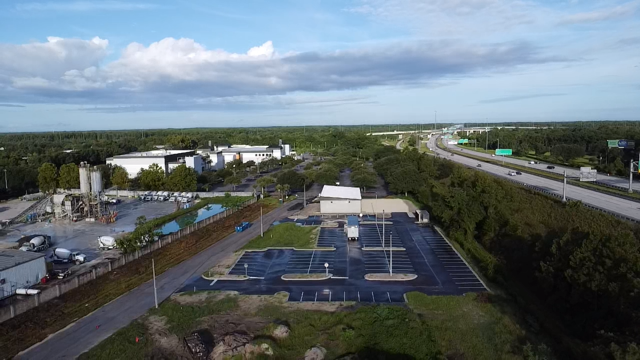 Aerial view of a parking lot near a highway with overpasses, surrounded by trees and industrial buildings under a partly cloudy sky.