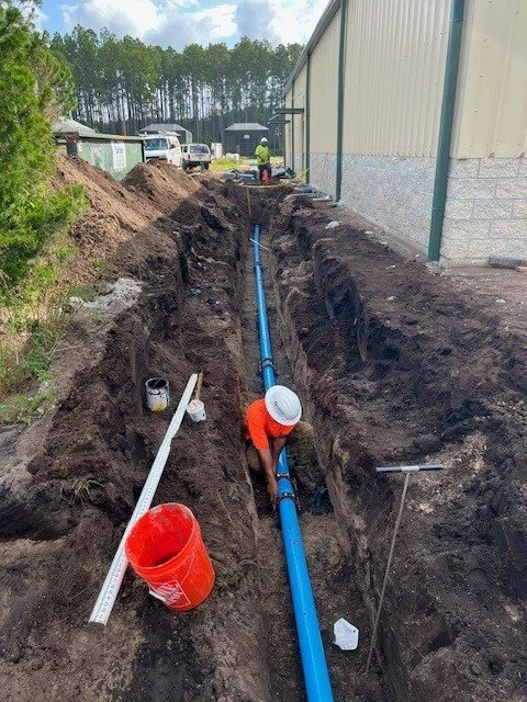 A construction worker installing a blue underground pipe in a trench outside a large building, with other workers and equipment visible in the background.