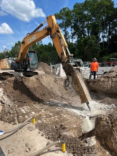An excavator with a hydraulic breaker attachment breaking up concrete in a construction site, with a worker in an orange shirt observing.