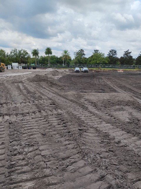 A dirt construction site with tire tracks, two cars in the distance, palm trees, and a cloudy sky.