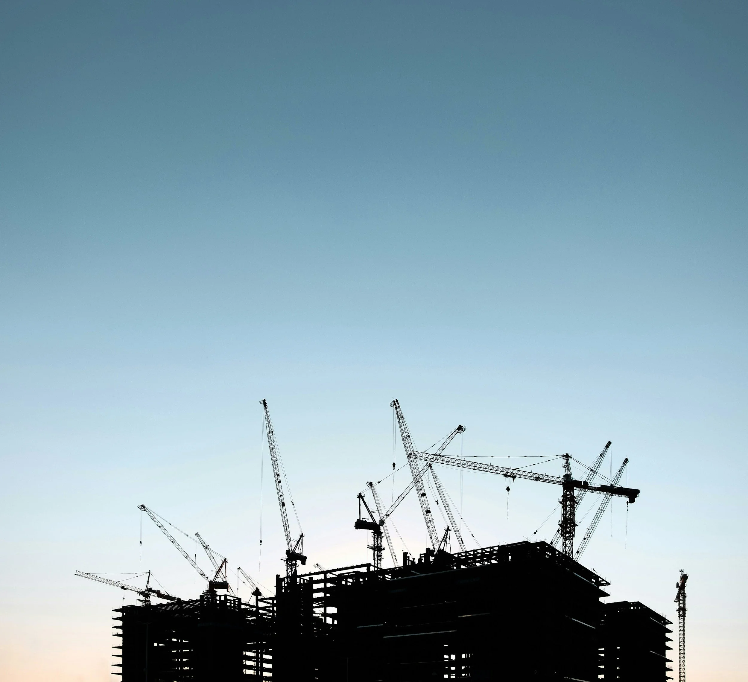Silhouette of a high-rise building under construction with multiple cranes against a blue sky at sunset.