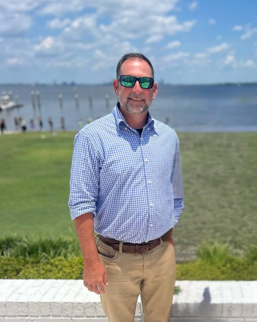 A man standing outdoors near a body of water with a grassy area in front of him. He is wearing sunglasses, a blue checkered shirt, and khaki pants, smiling at the camera. The background includes boats, a pier, and cloudy blue sky.