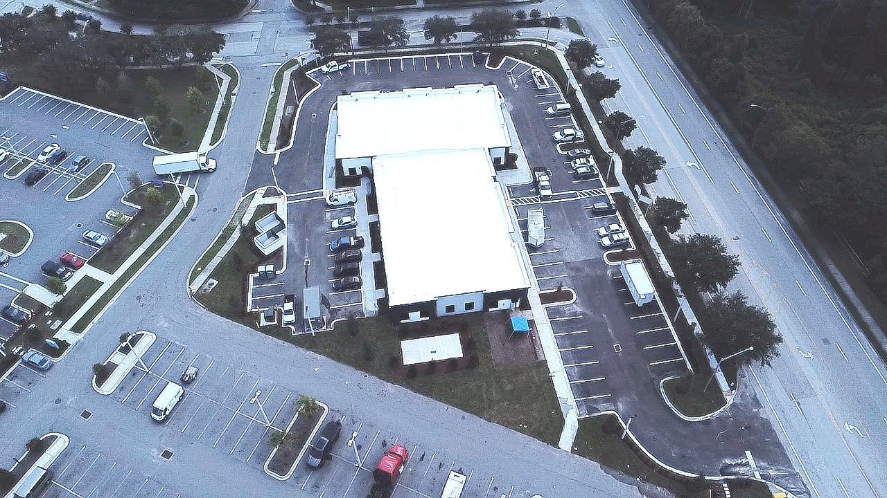 An aerial view of a commercial building with a parking lot, surrounded by trees, and adjacent to a road with multiple lanes.
