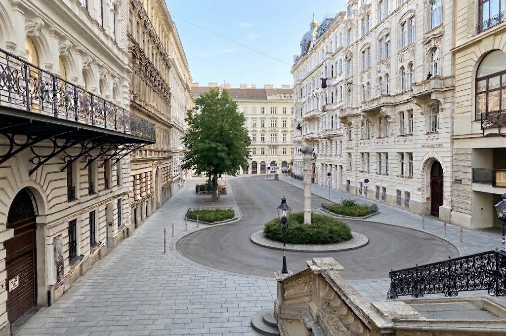 Empty streets lined with old, multi-story white buildings and a central fountain featuring a statue in a historic square in a European city.