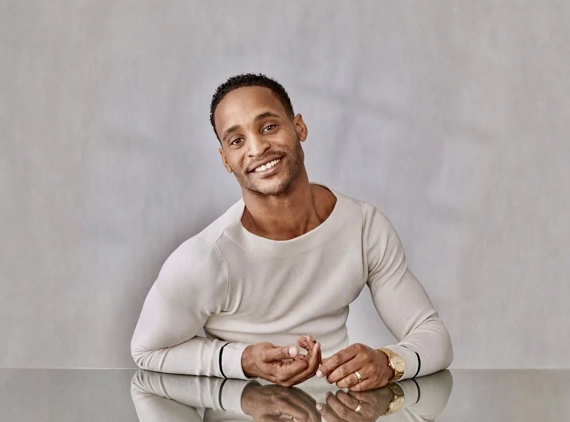 A smiling man is sitting at a table in front of a plain wall. He is wearing a light gray long-sleeved shirt, a wristwatch, and a ring.