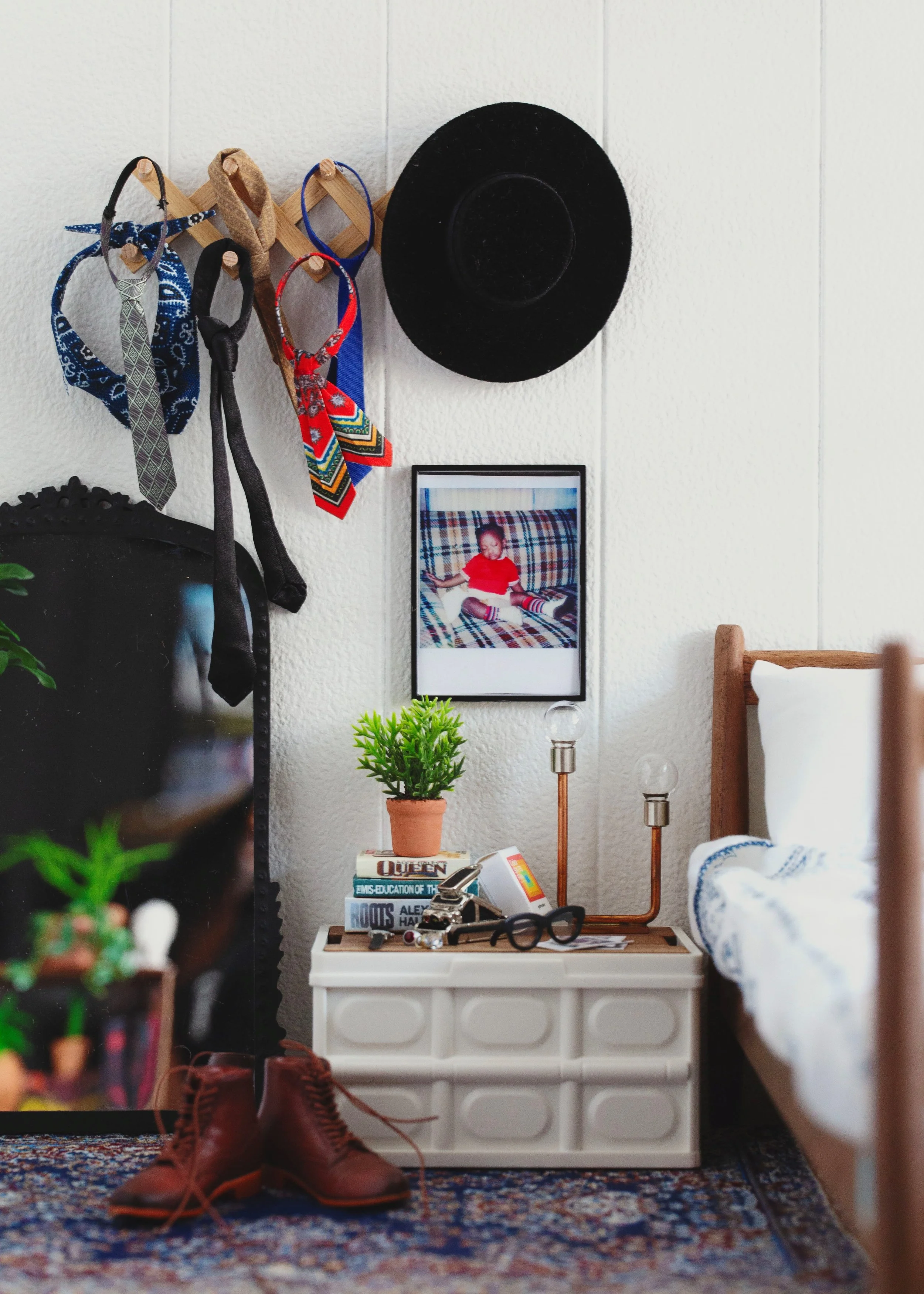 Bedroom corner with a white nightstand, a photo of a child on the wall, a potted plant, books, glasses, and a pair of brown boots on the floor. Several colorful bandanas and a black hat hang on the wall.