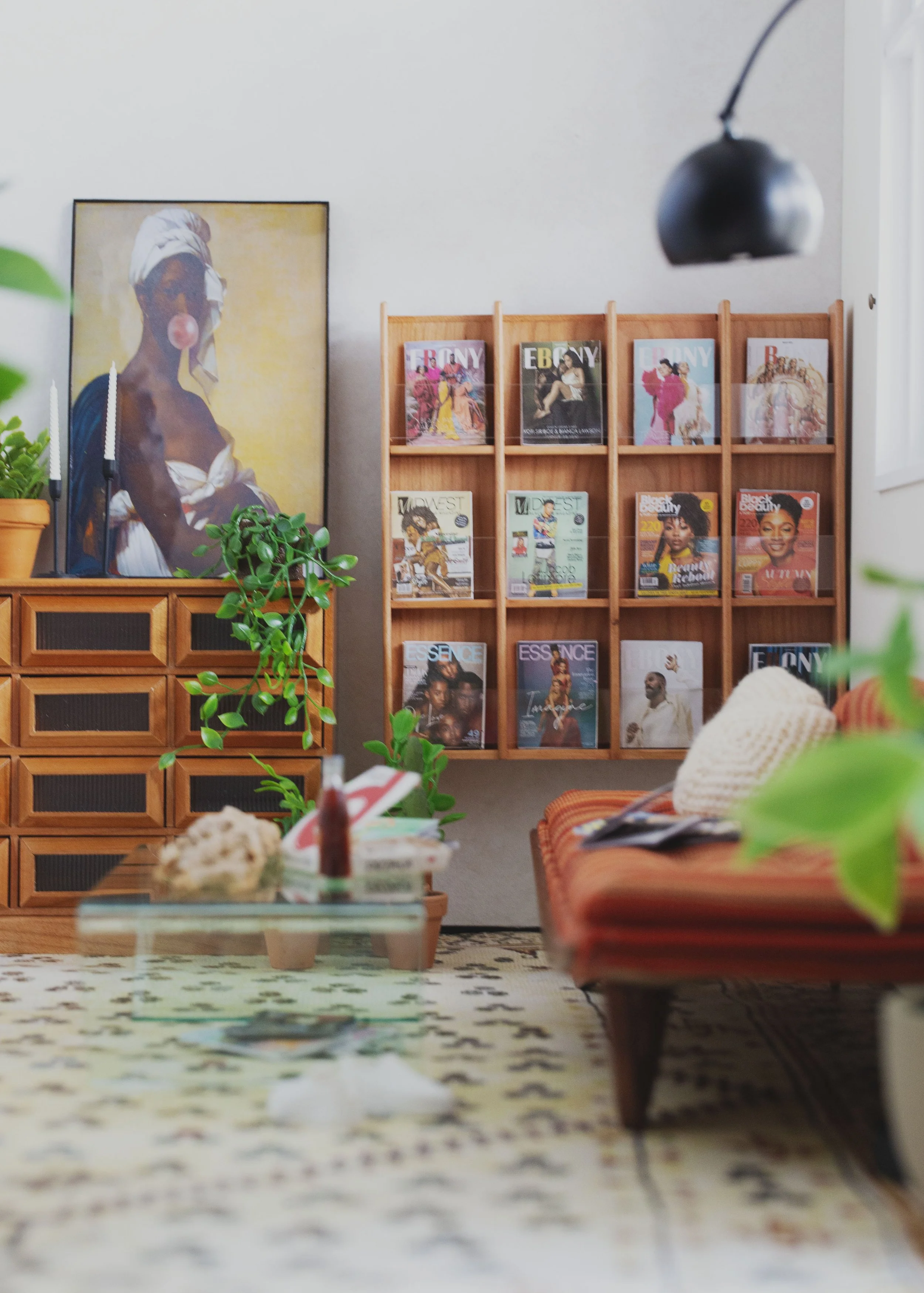 Living room with a wooden magazine rack filled with fashion magazines, a painting of a woman with a towel on her head and bubblegum, potted plants, a glass coffee table with decor, an orange couch with pillows, and a black hanging lamp.