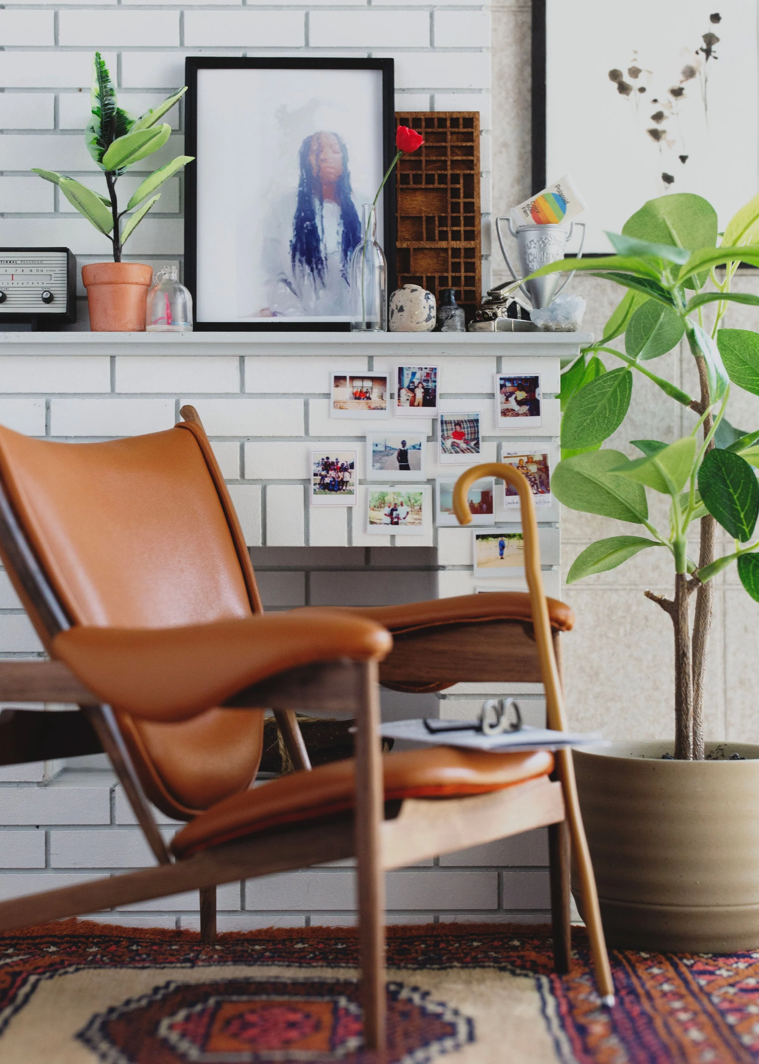 A cozy interior scene featuring a white brick fireplace with a photo collage, potted plants, and decorative items on the mantel. In front, a tan leather and wood lounge chair with a wooden cane walking stick resting on it. The floor has a colorful patterned rug.