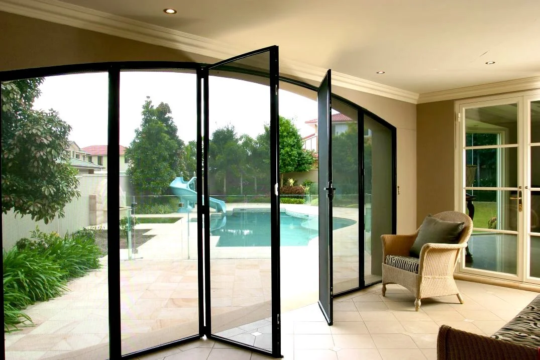 Interior view of a living room with a folding glass door open to a backyard with a swimming pool and a waterslide, outdoor trees, and neighbor houses.