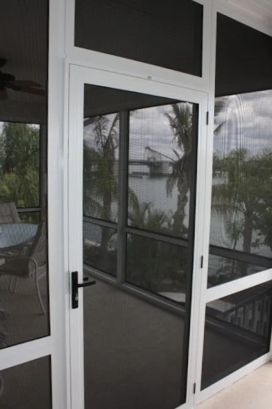 View of a screened-in porch with a white door and black handle, overlooking palm trees and a body of water outside.