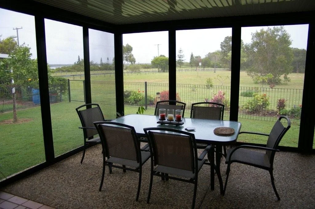 Screened porch with a black metal table and six chairs, candles on a tray, overlooking a grassy yard and garden with trees and a fence.