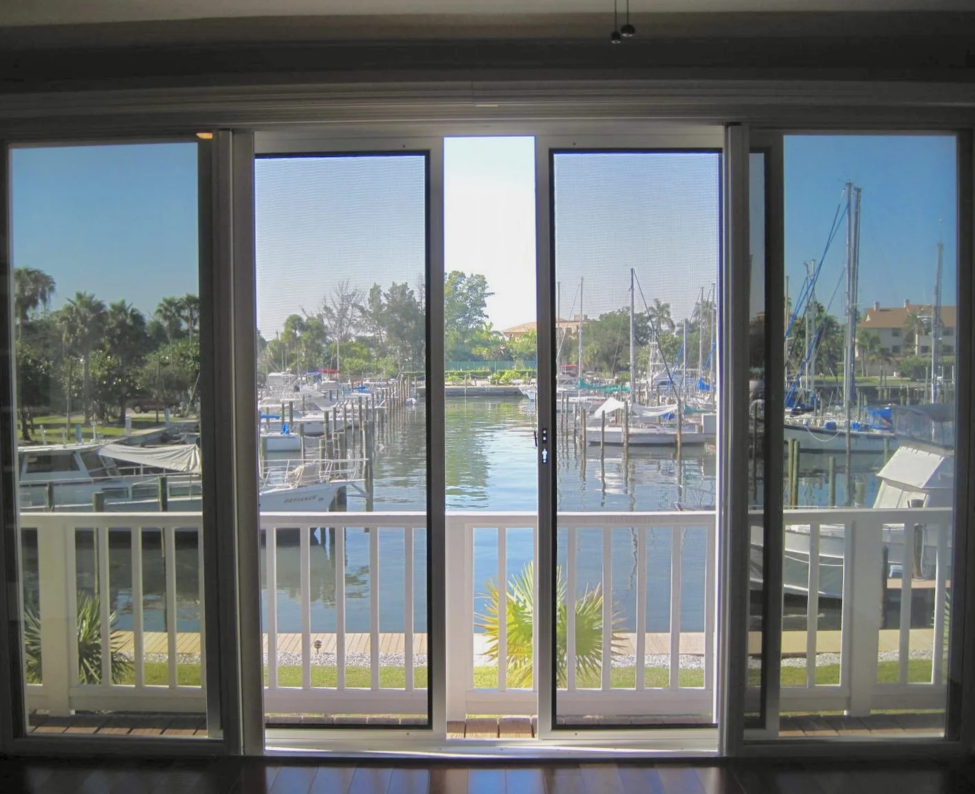 View of a marina with sailboats and motorboats docked, seen through sliding glass doors from inside a room with a balcony.
