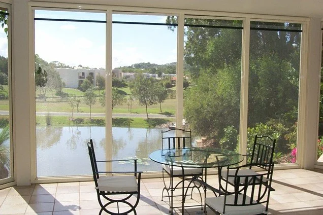 Sunlit dining area with a glass-topped table and four black metal chairs in front of large glass windows overlooking a pond and greenery.