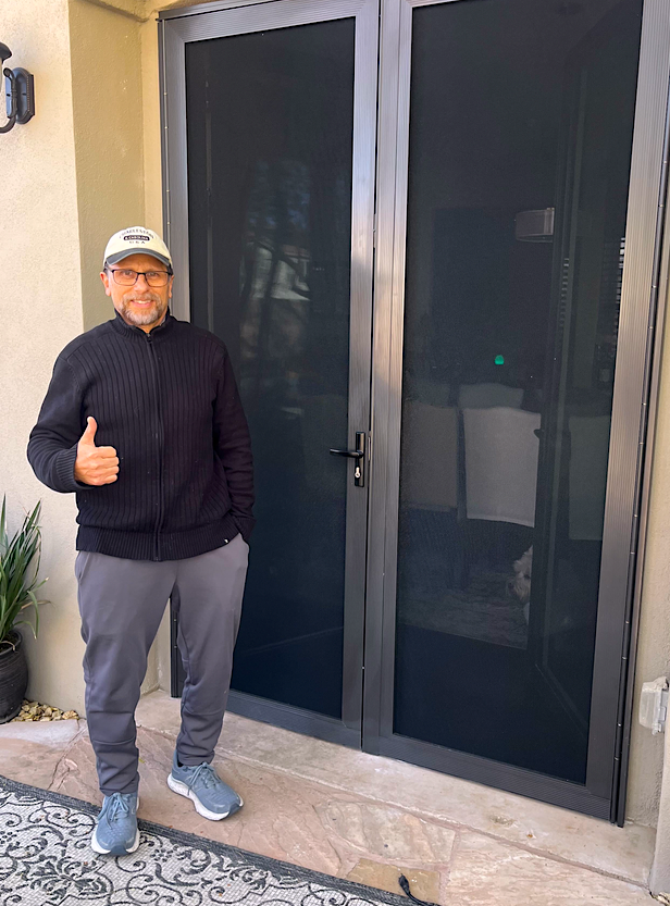 A man smiling and giving a thumbs-up gesture while standing outside near a black framed glass door, wearing glasses, a white cap, black jacket, gray pants, and sneakers.