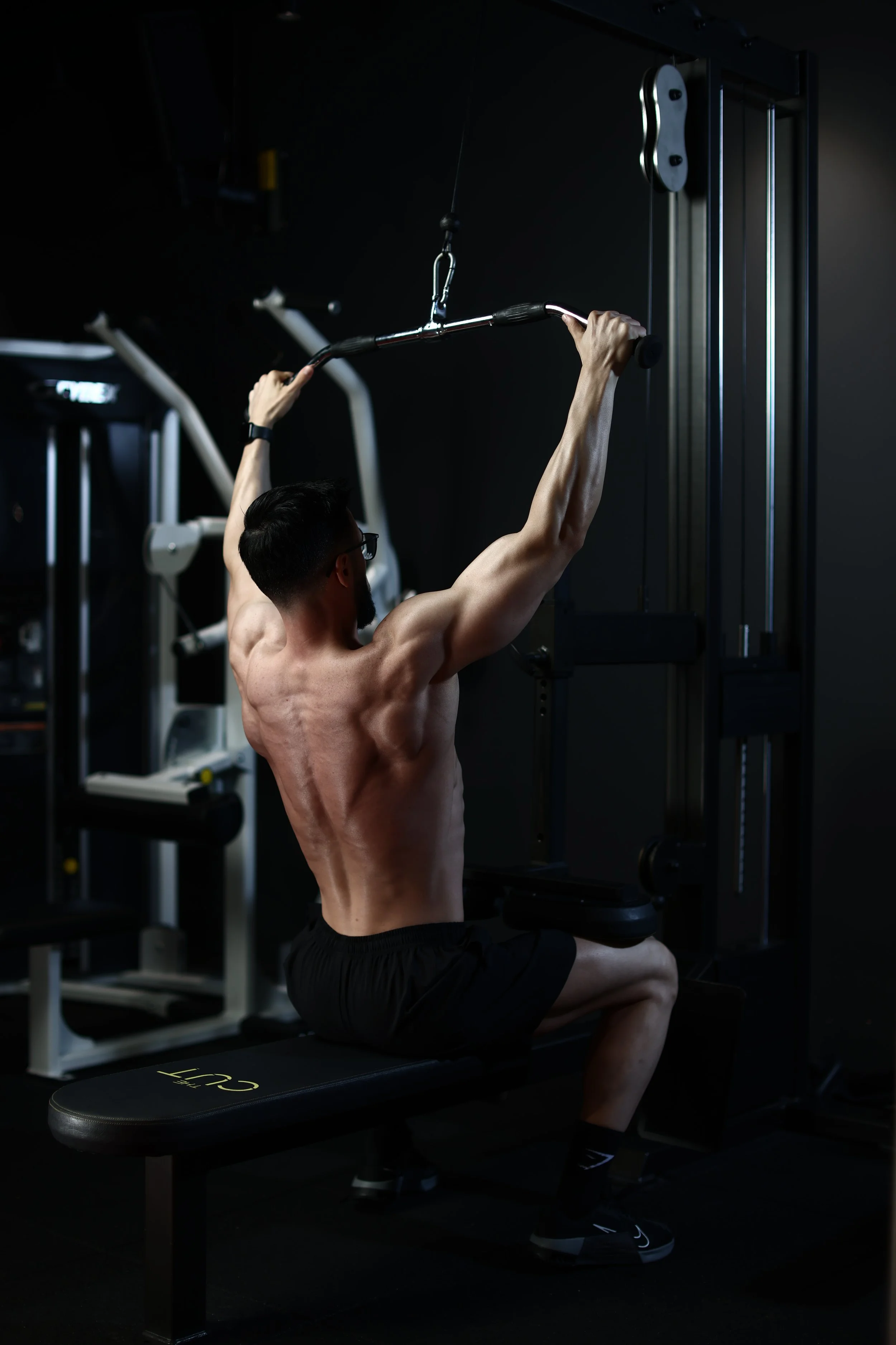 A shirtless man with glasses sitting on a workout bench, using a lat pulldown machine in a dark gym.