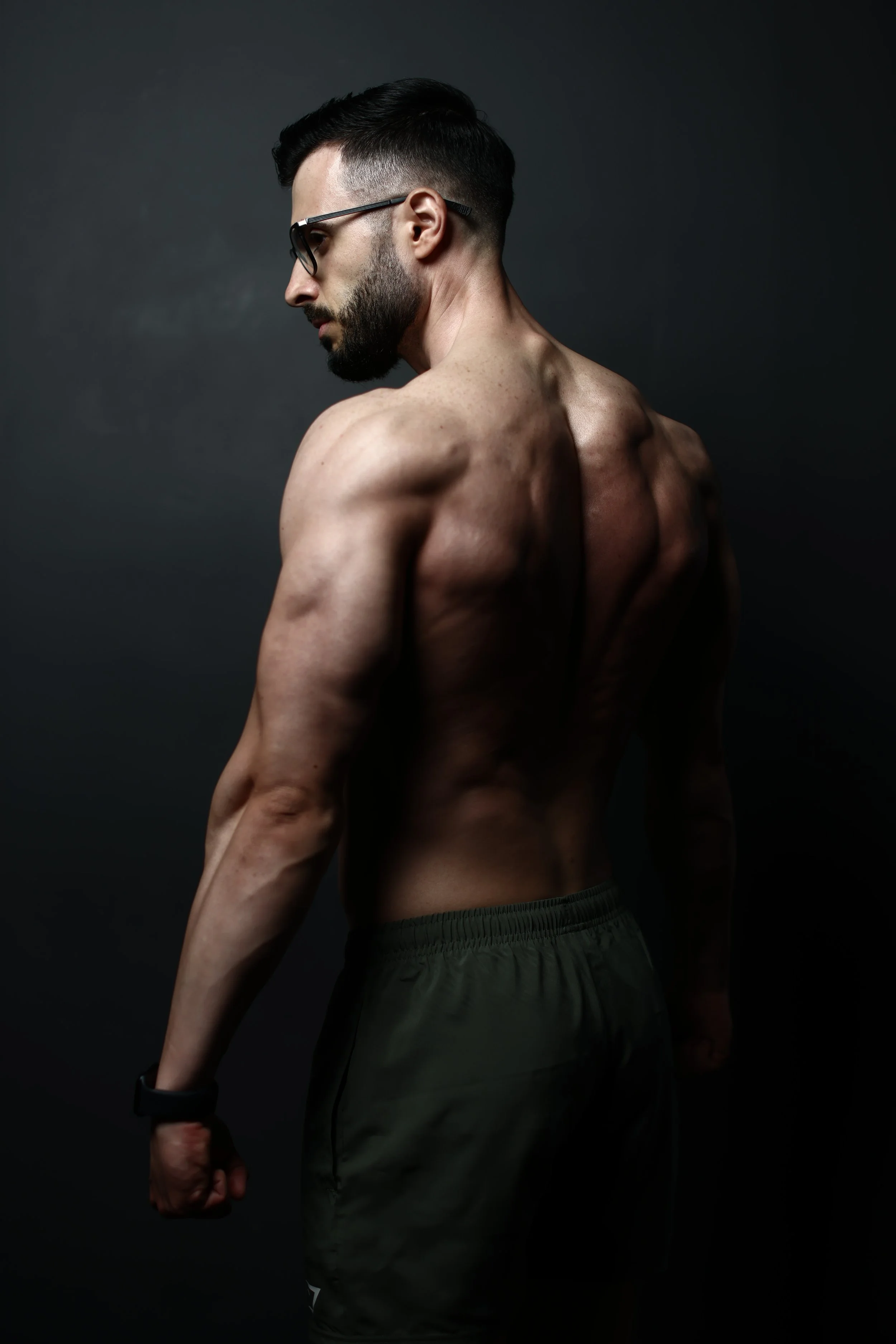 A shirtless man with a beard and glasses looking down, with a dark background.
