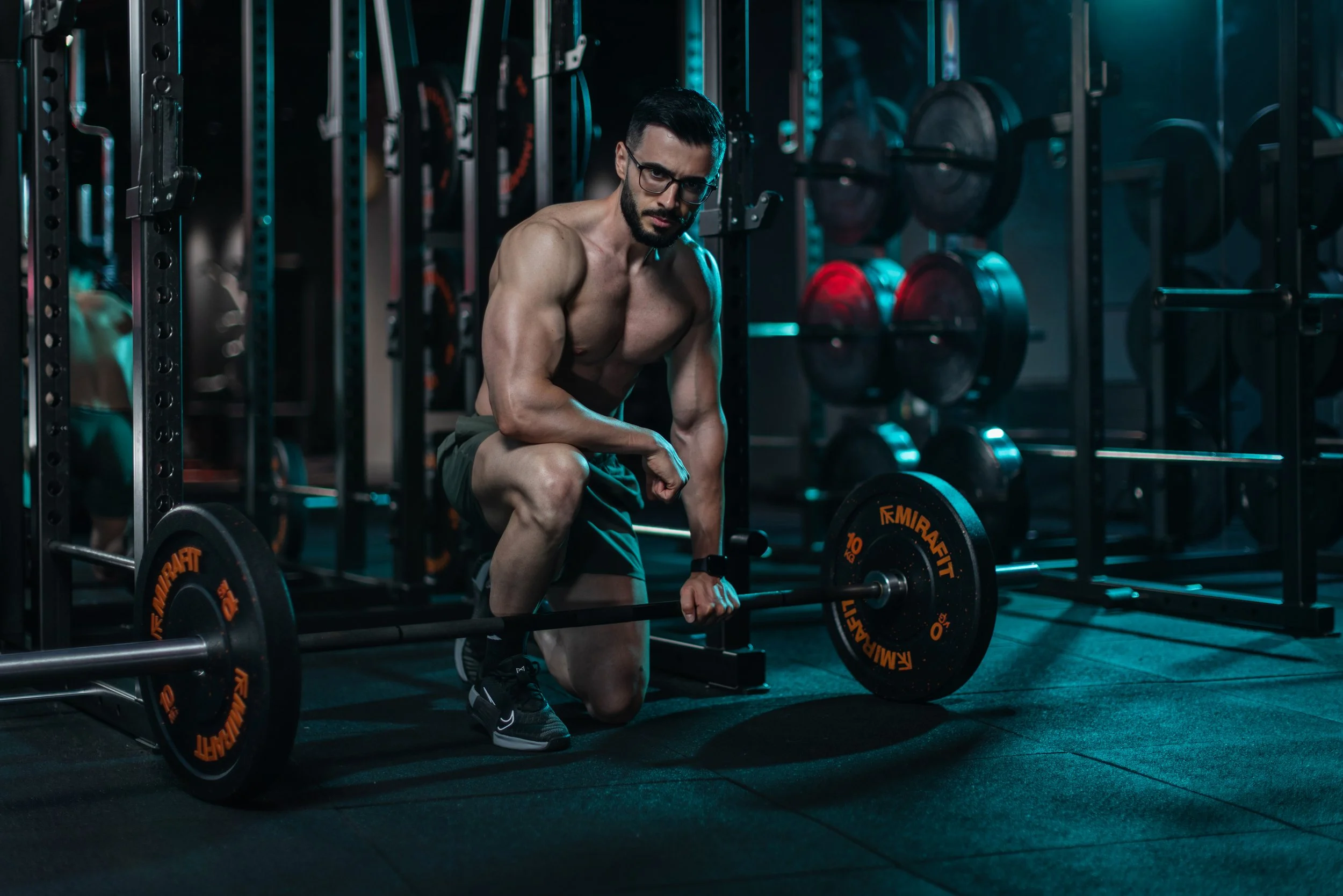 A muscular man with glasses, kneeling on the gym floor, holding a barbell with weights labeled 'Emiragit', in a dimly lit gym with weight racks in the background.