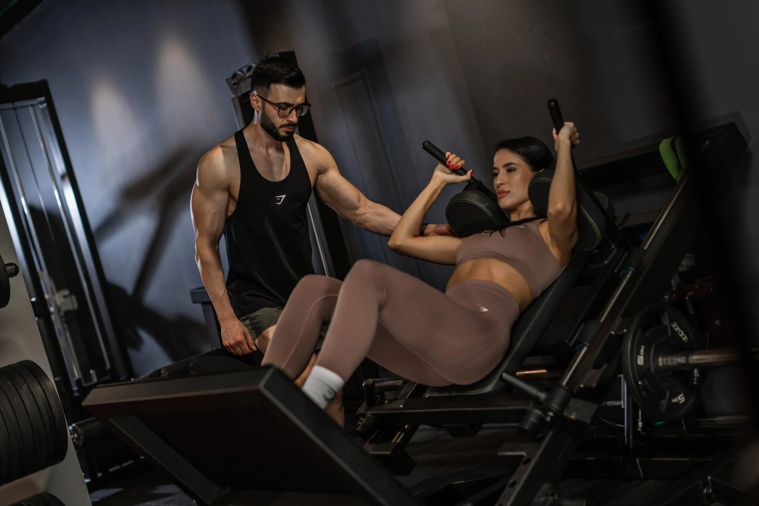 A woman in workout clothes lying on a leg press machine while a trainer assists her during exercise.