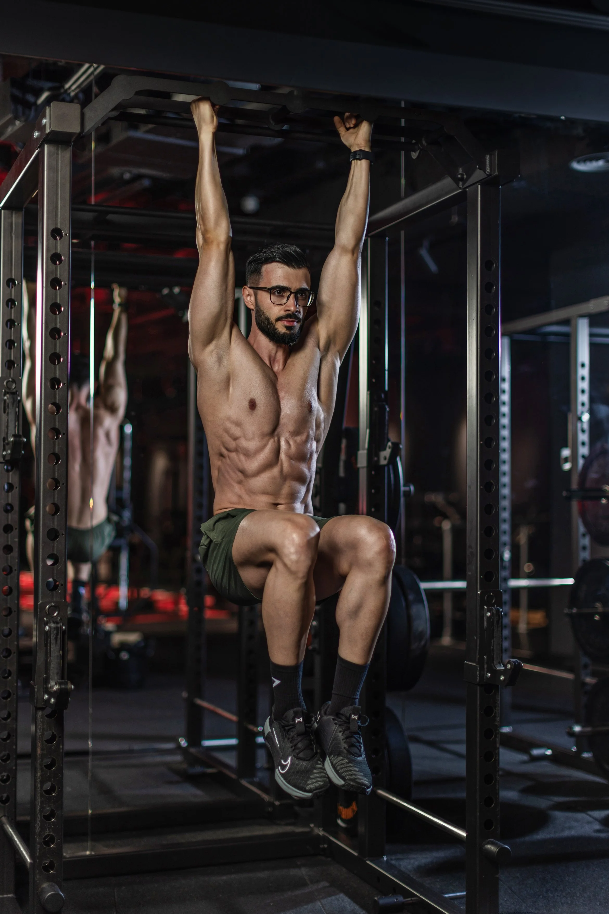 A shirtless man with glasses and a beard is performing a hanging leg raise exercise on a workout cage in a gym. He is wearing dark shorts, black socks, and athletic shoes, and is holding onto the top of the cage with both hands, with another person doing the same exercise in the background.