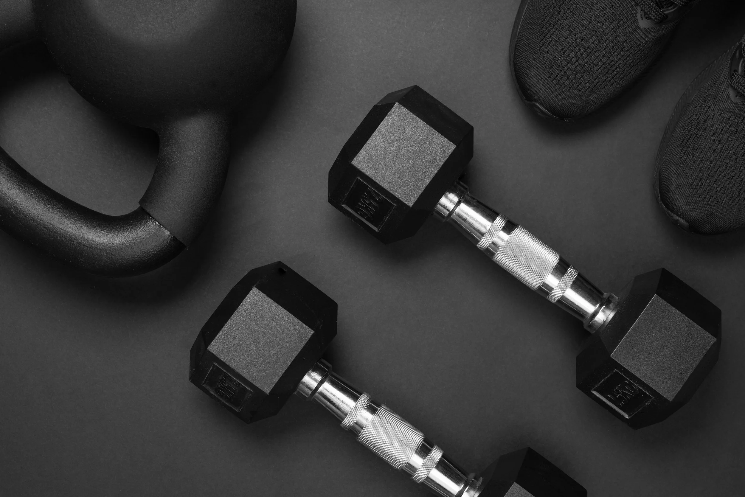 Black kettlebell, pair of black dumbbells, and a pair of black athletic shoes on a black surface.