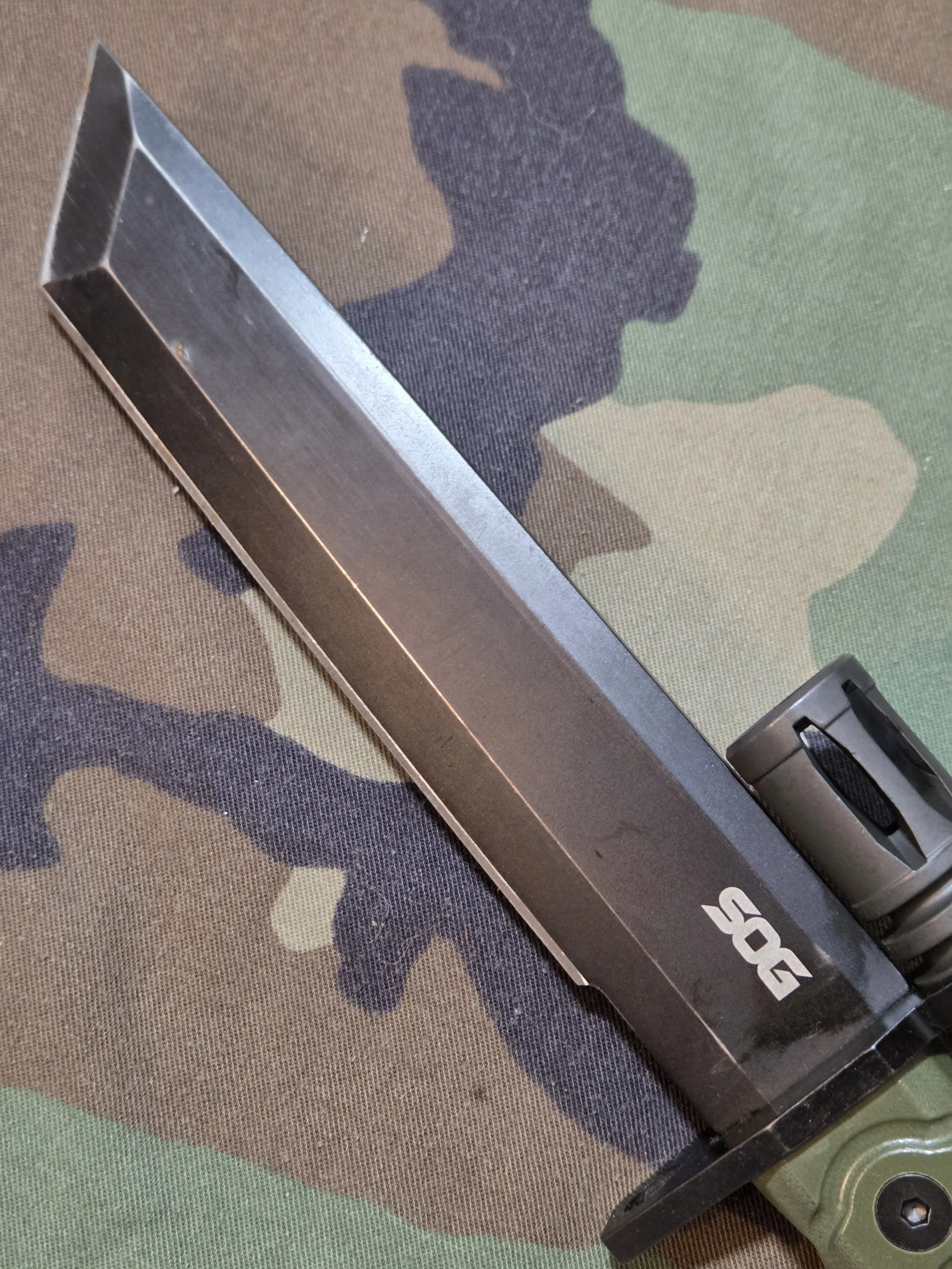 black tanto knife blade with a woodland camouflage background. SOG on the blade with OD green G10 grip under an A2 birdcage flash hider.