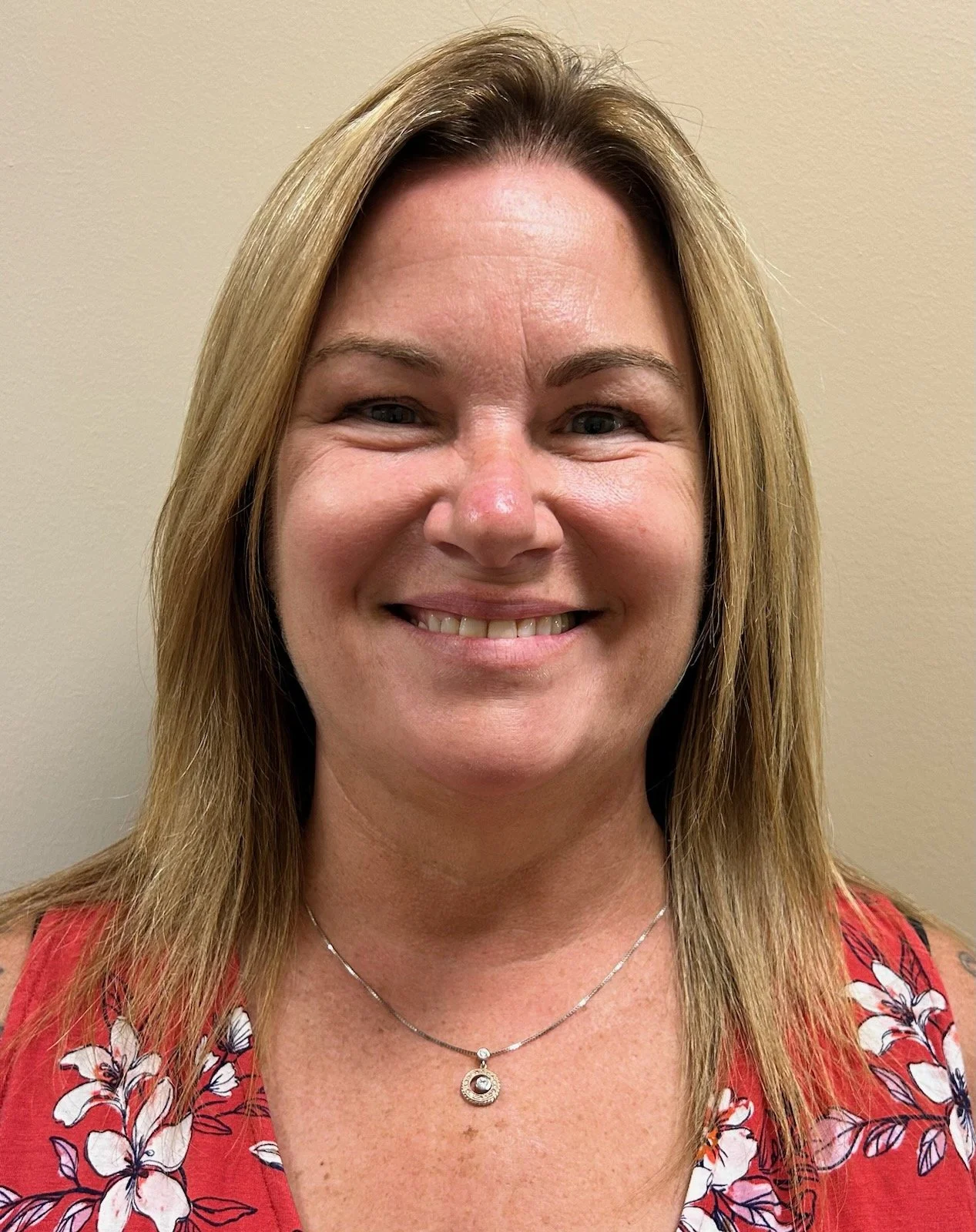 A woman with shoulder-length blonde hair smiling in front of a beige wall, wearing a red floral top and a silver necklace.