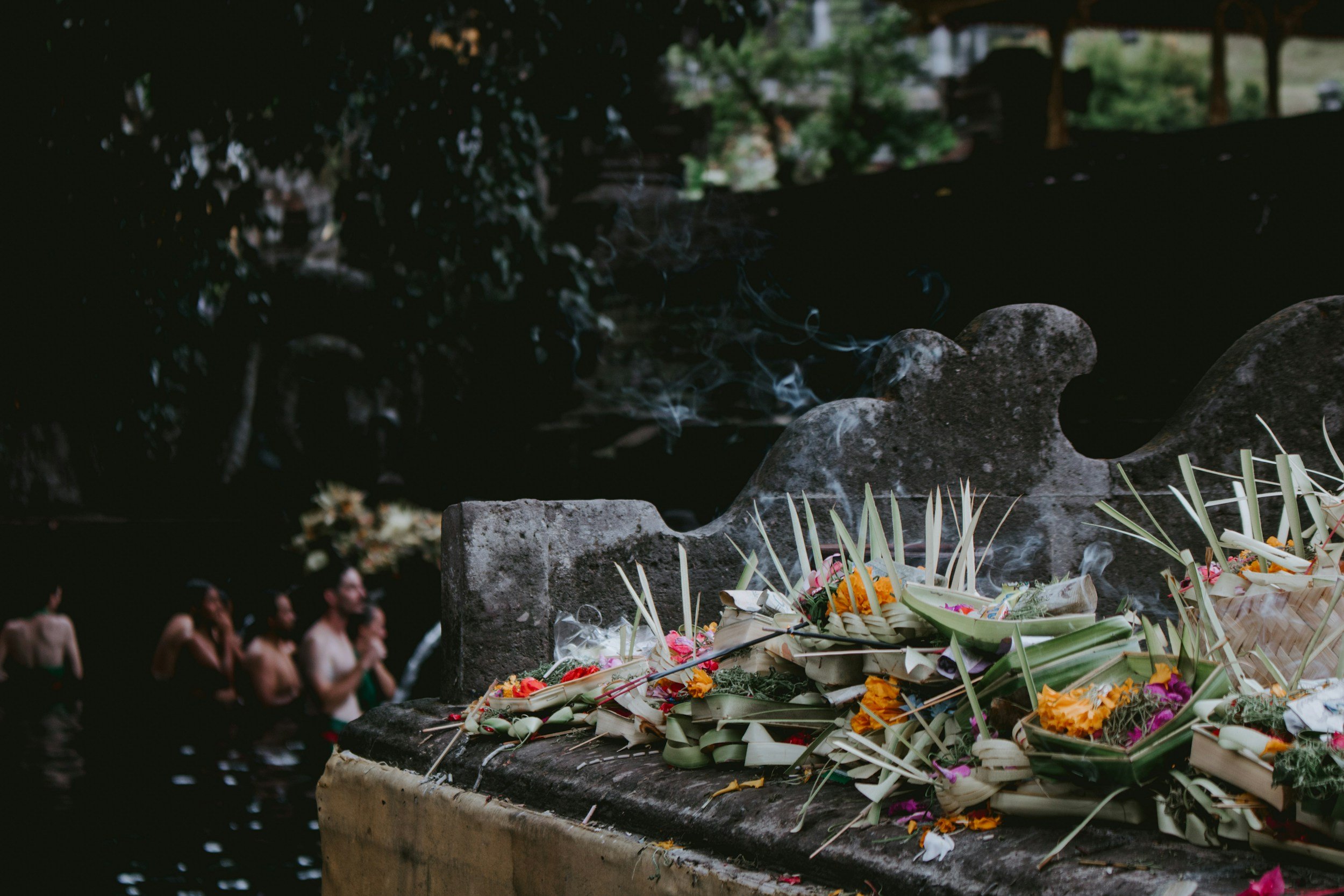 Flowers, incense, and offerings placed on a stone platform in a traditional outdoor setting. In the background, a group of people stand in water, partially submerged, participating in a ritual or ceremony.