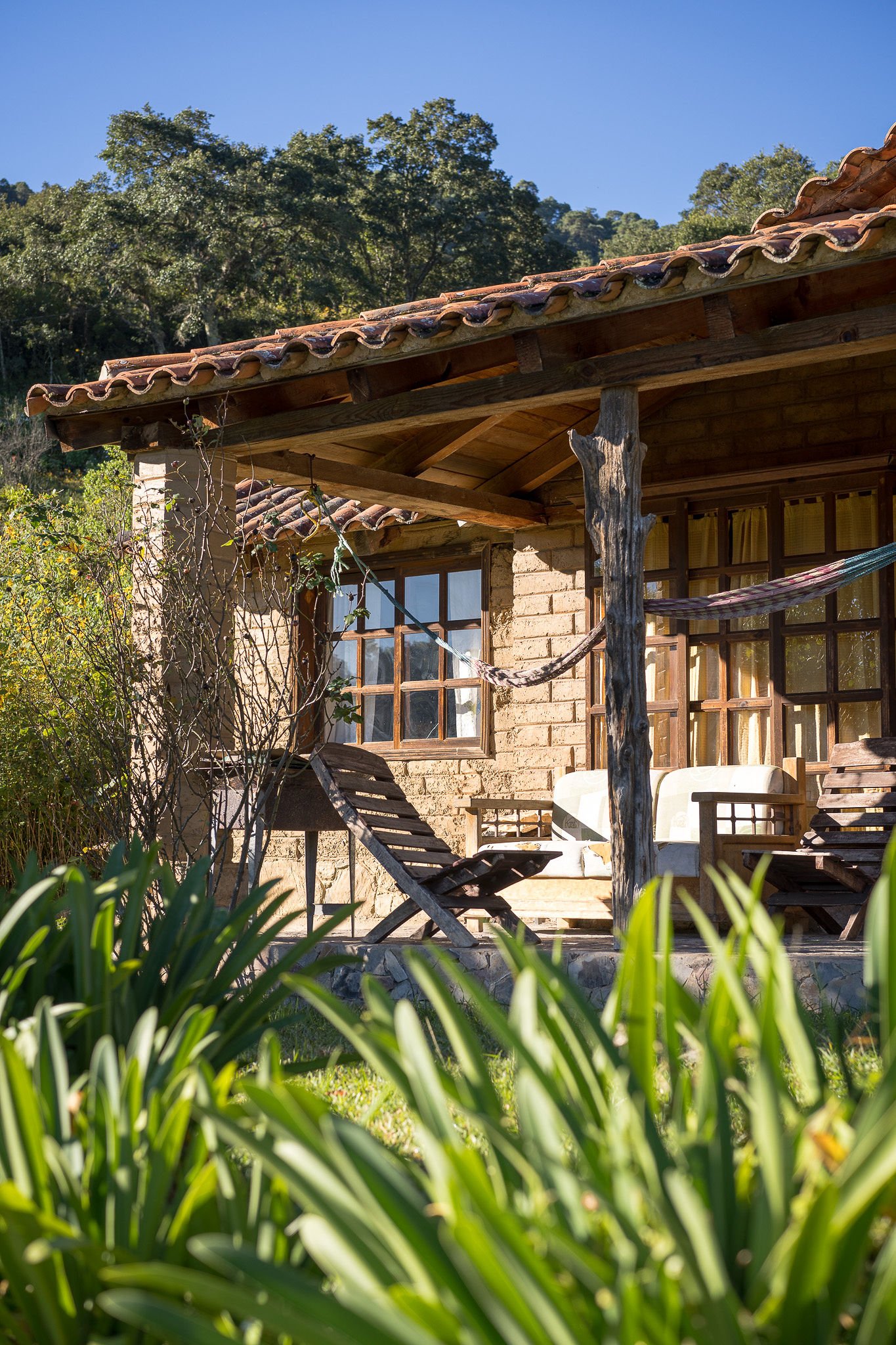 A rustic house with a wooden porch, surrounded by greenery, with outdoor furniture including lounge chairs and a hammock, under a clear blue sky.