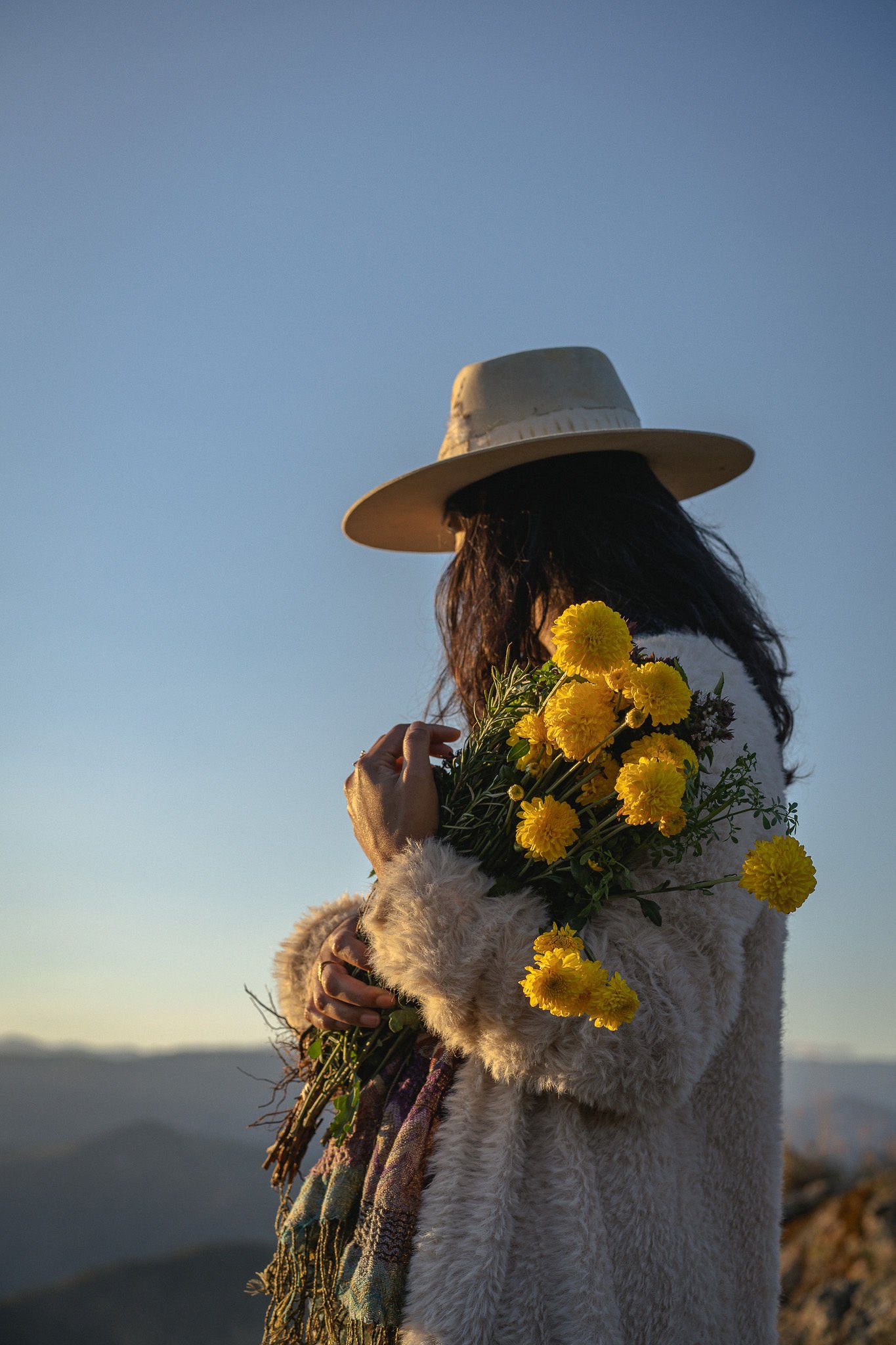 Woman holding yellow flowers, wearing a beige hat and a fluffy coat, standing outdoors during sunset.
