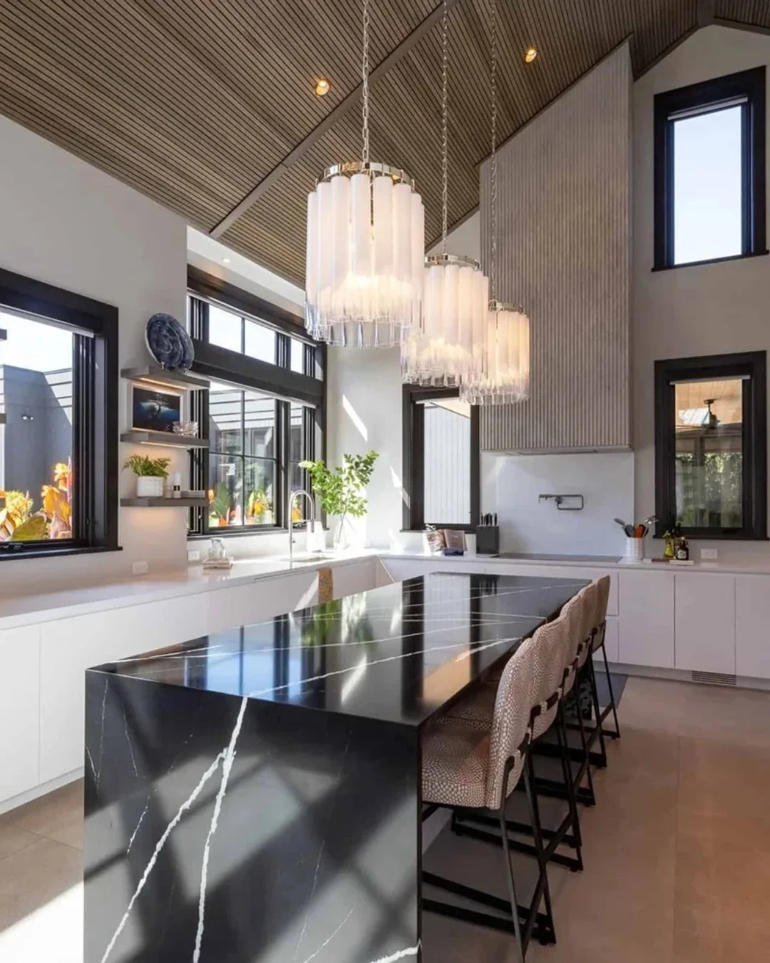 A kitchen that makes a statement. The dramatic waterfall island sets the tone, grounded by clean white cabinetry and framed by natural light from every angle. Vertical paneling and warm wood ceilings add texture above, while layered lighting brings i