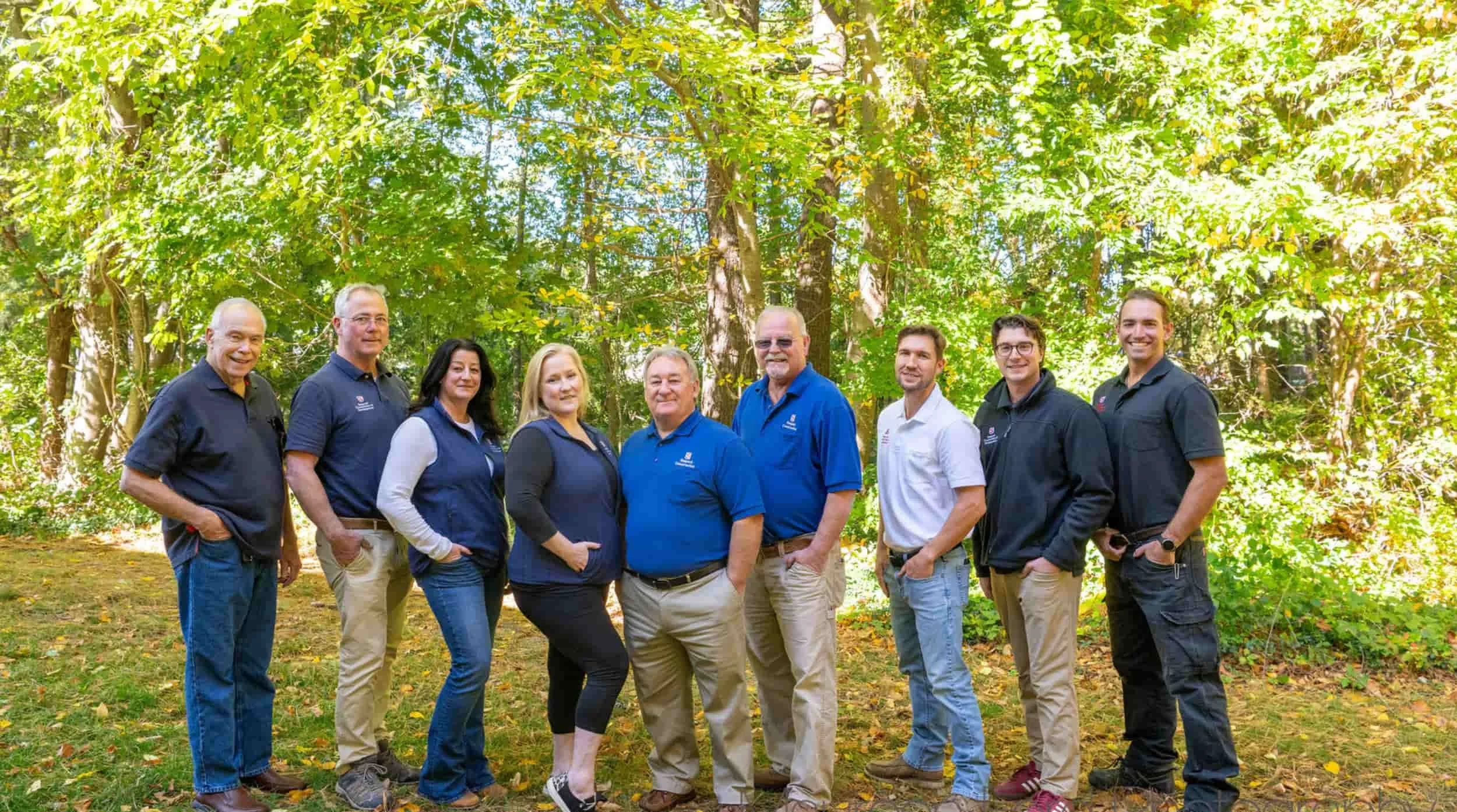 Group of eleven people standing outdoors in a wooded area, smiling for the photo.