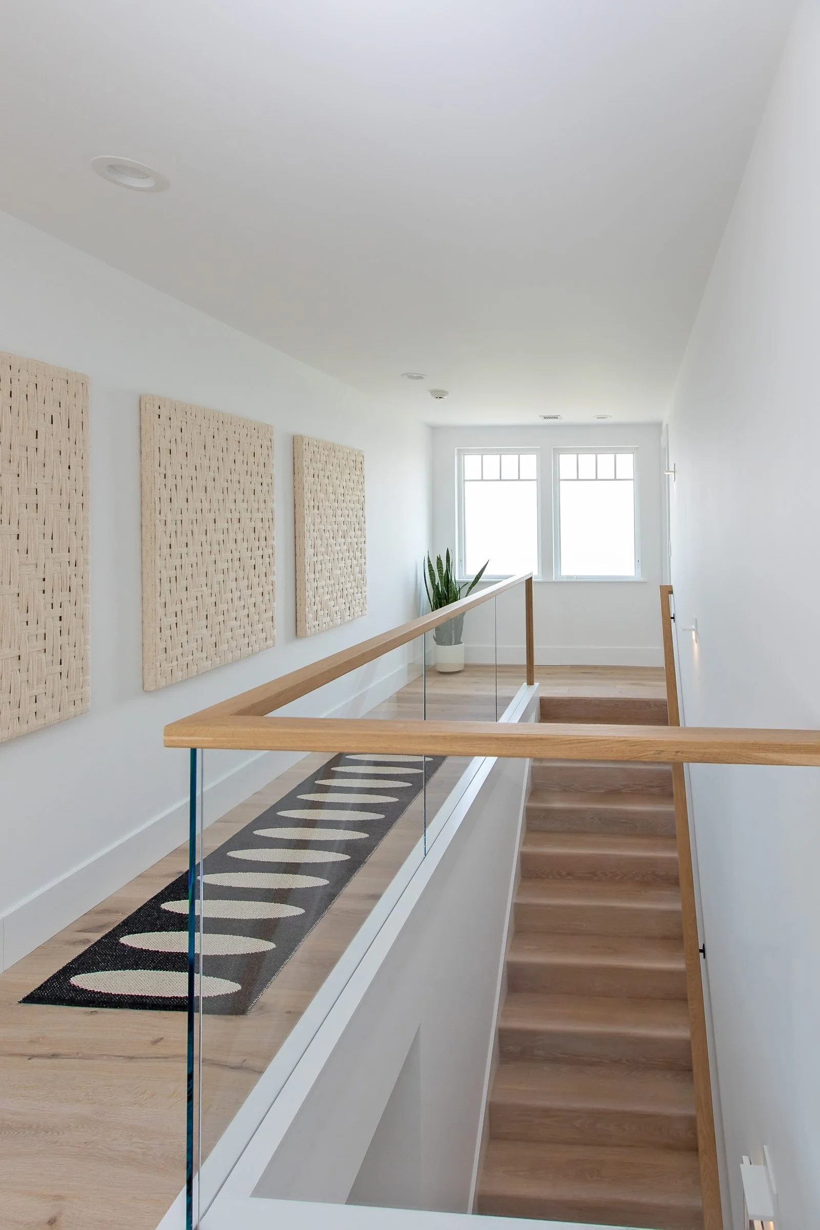 Bright hallway with wooden stairs, glass railing, and three woven wall hangings, featuring a potted plant near the window at the end.